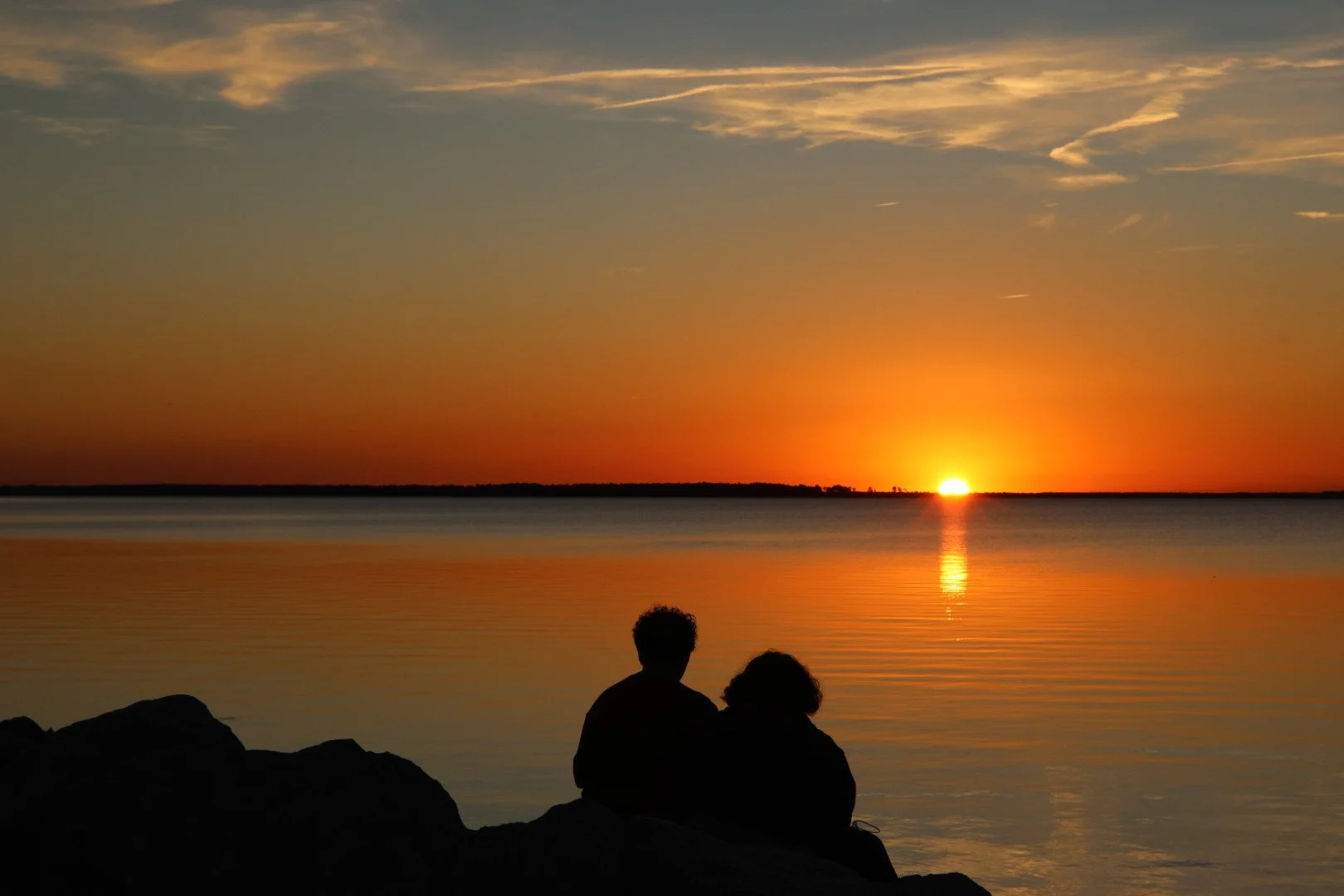 Silhouettes of two people sitting on rocks by a calm body of water, watching a sunset with orange and yellow hues reflected on the water, with a few clouds in the sky.