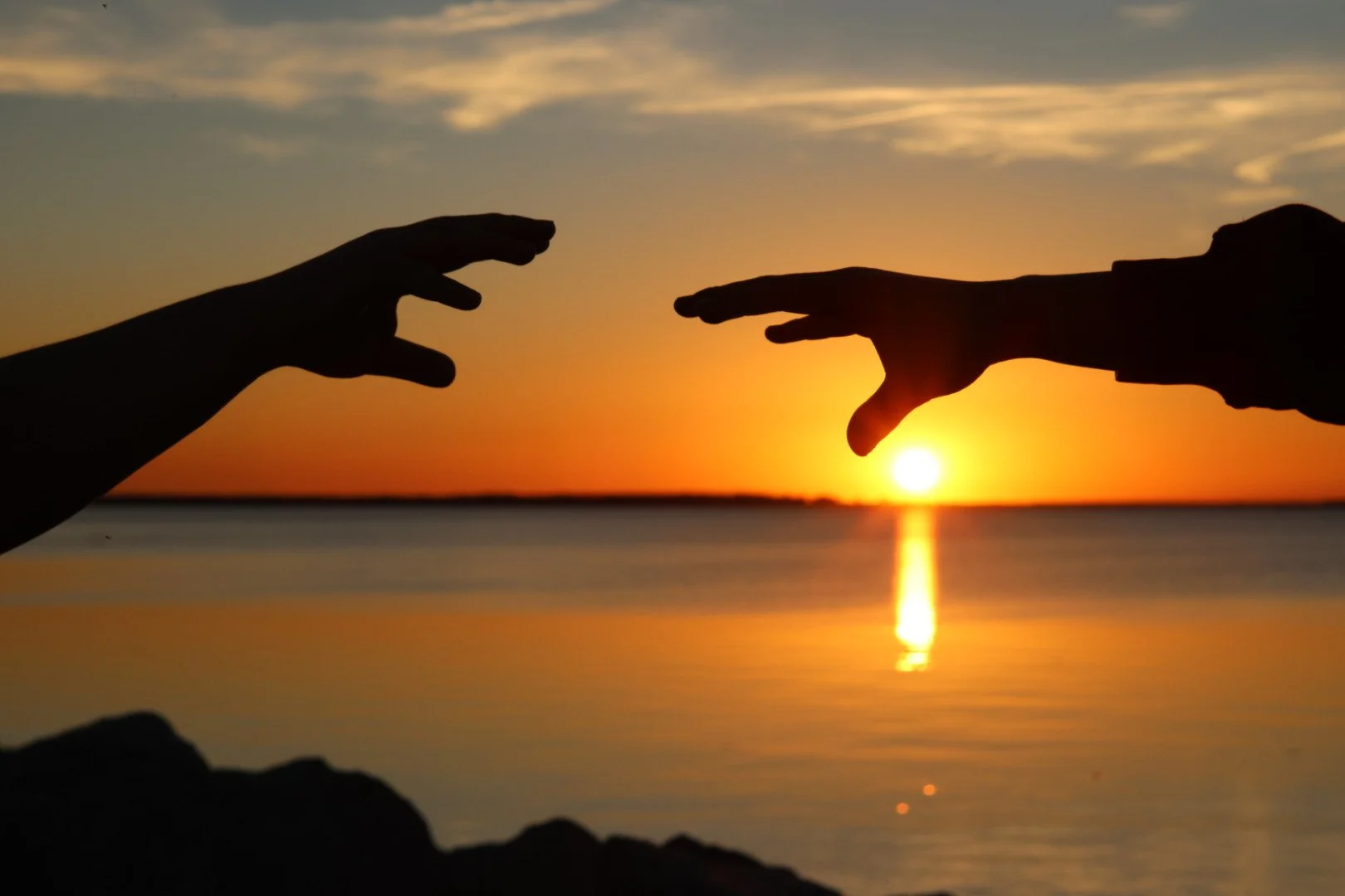 Silhouettes of two hands reaching towards each other with a sunset over water in the background.