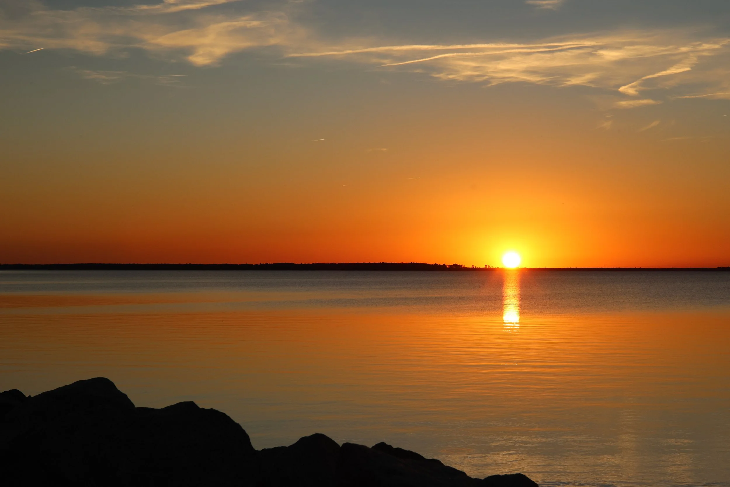 Sunset over a calm body of water with a silhouette of rocks in the foreground and a horizon with some trees in the distance.