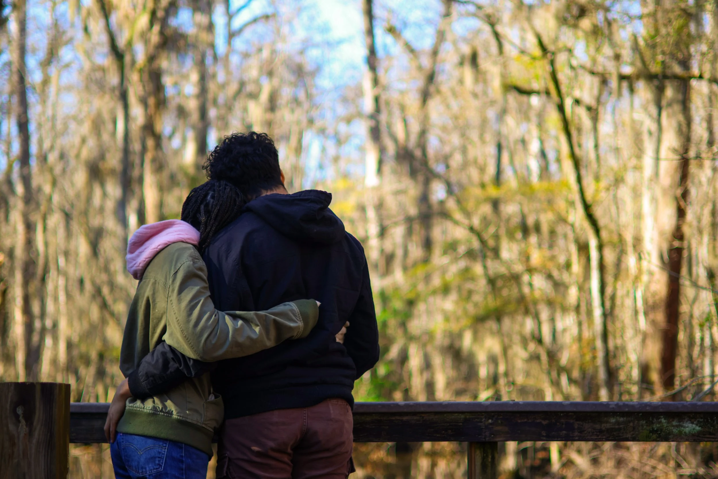 Two people with dark curly hair, one wearing a black hoodie and the other a green jacket with a pink hood, are standing closely together outside, leaning on a wooden railing in a wooded area with trees.