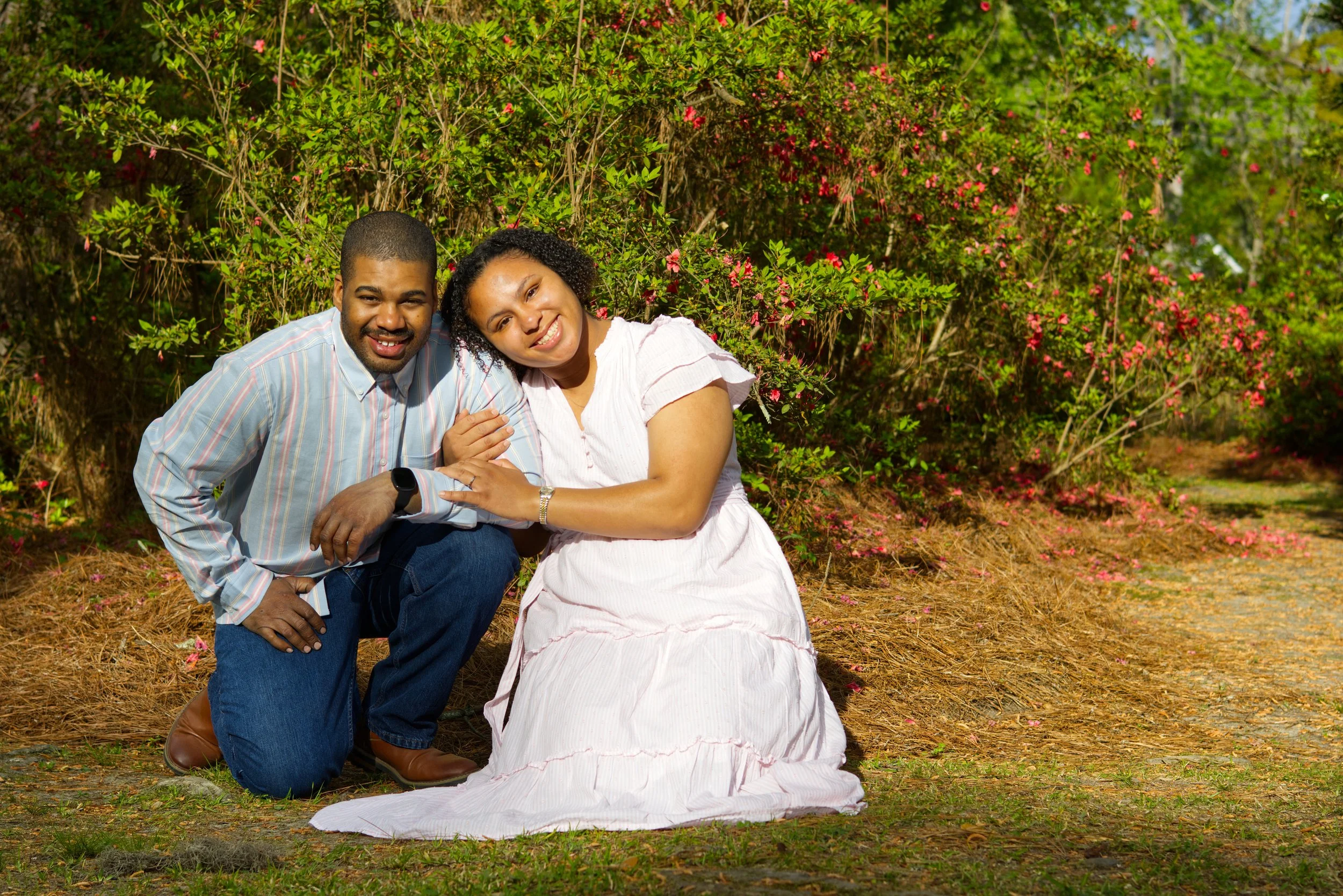 A smiling couple kneeling on the ground outdoors in front of flowering bushes, with the woman wearing a white dress and the man in a striped shirt.
