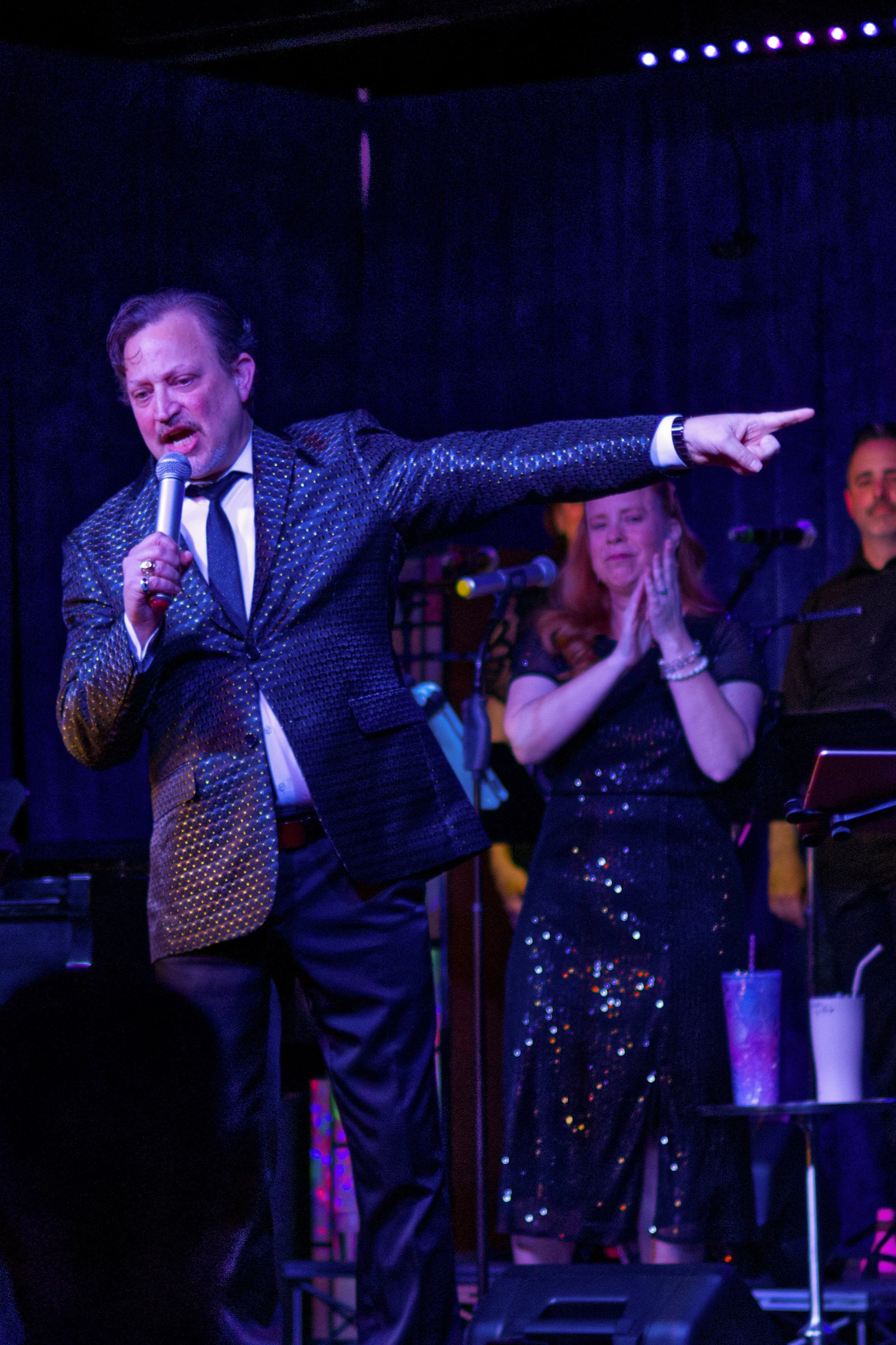 Man singing into a microphone on stage, pointing outward, with a woman behind him clapping and a man smiling in the background, all dressed in formal attire, performing at a concert or event.
