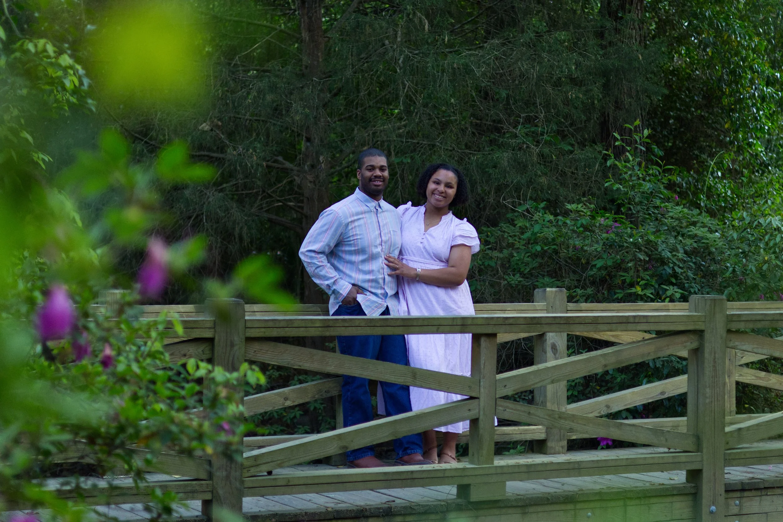 A smiling couple standing on a wooden bridge in a lush, green outdoor setting.