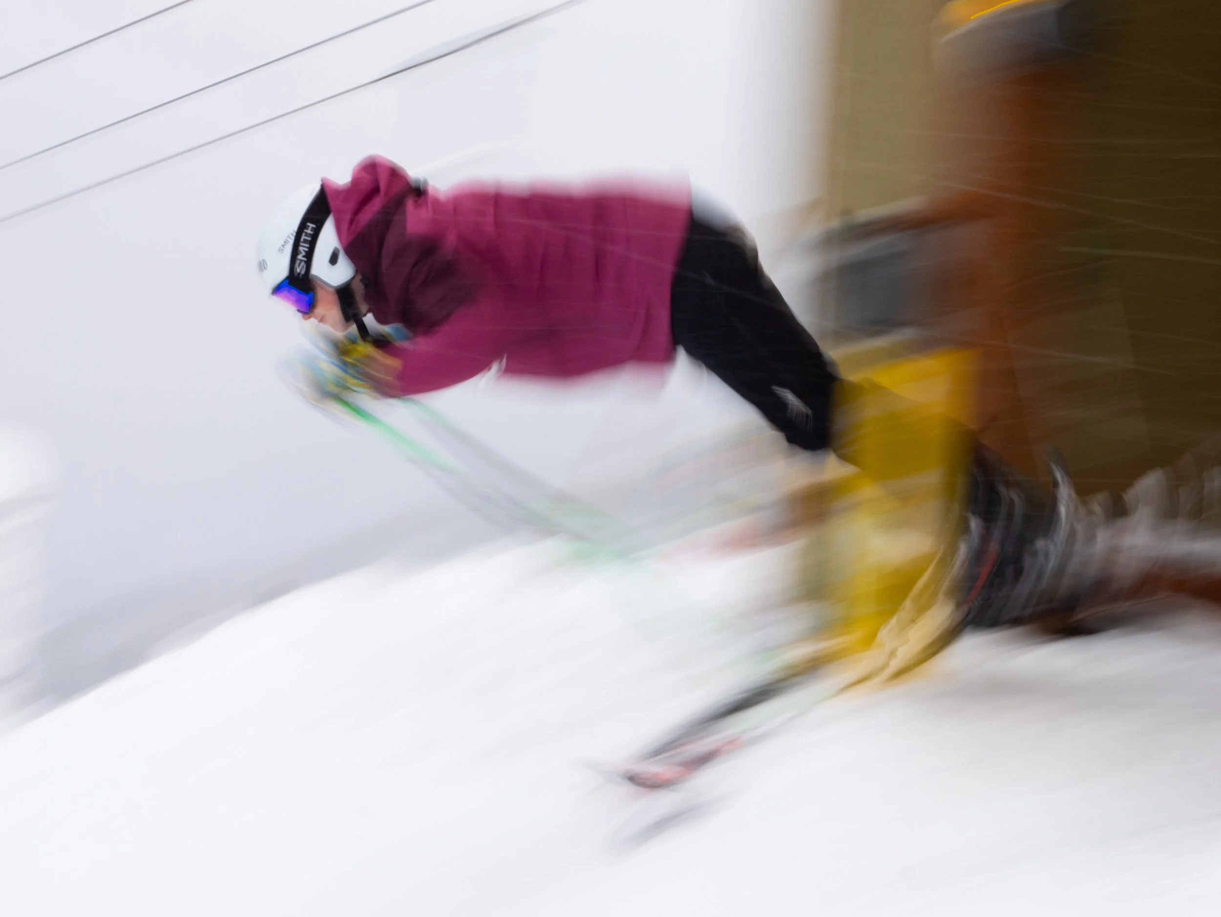 A person wearing a white helmet, maroon jacket, and black pants skiing or snowboarding on a snowy slope, holding ski poles.