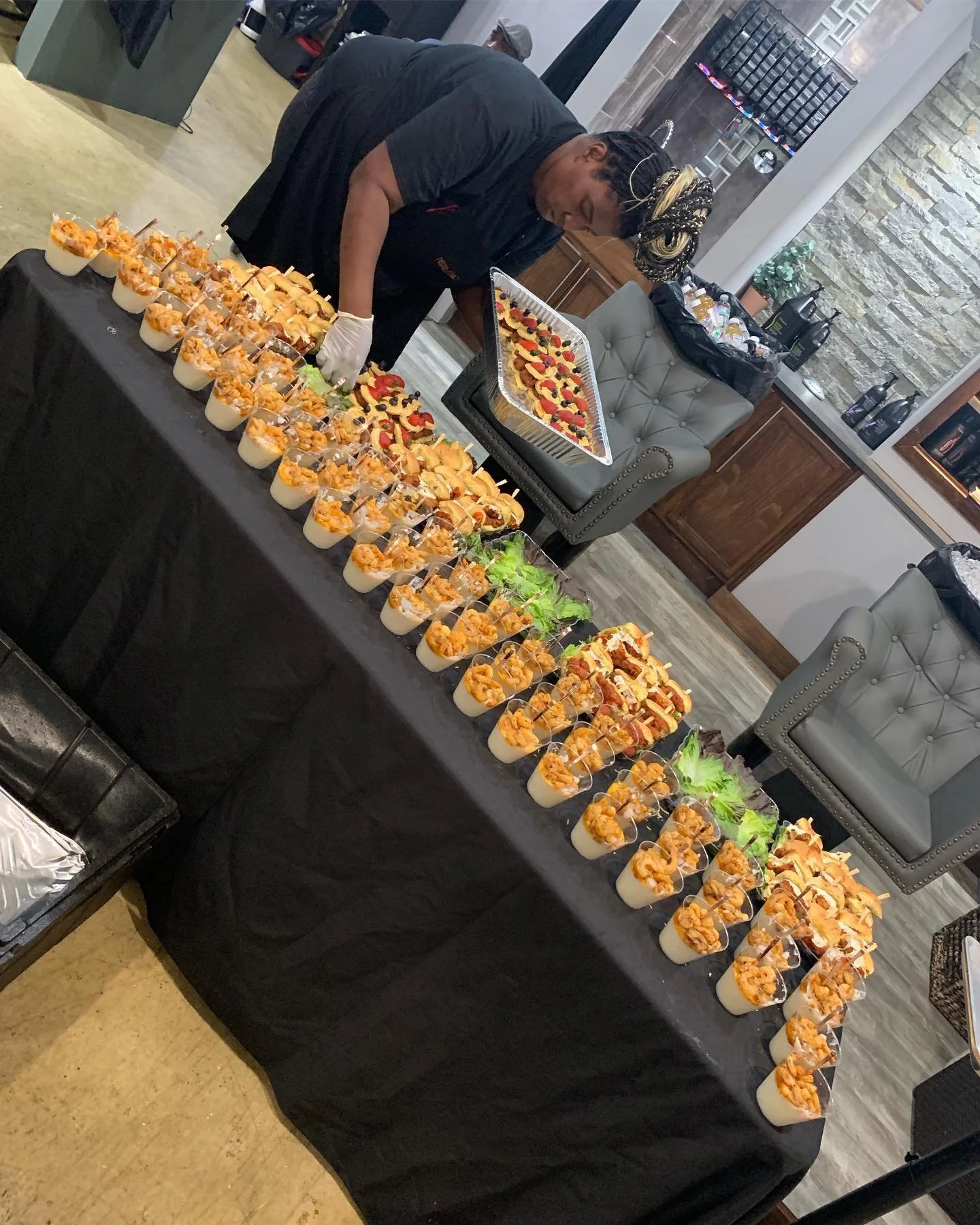 Boomer preparing a large black table filled with serving cups containing shrimp and grits, in a catering setting.