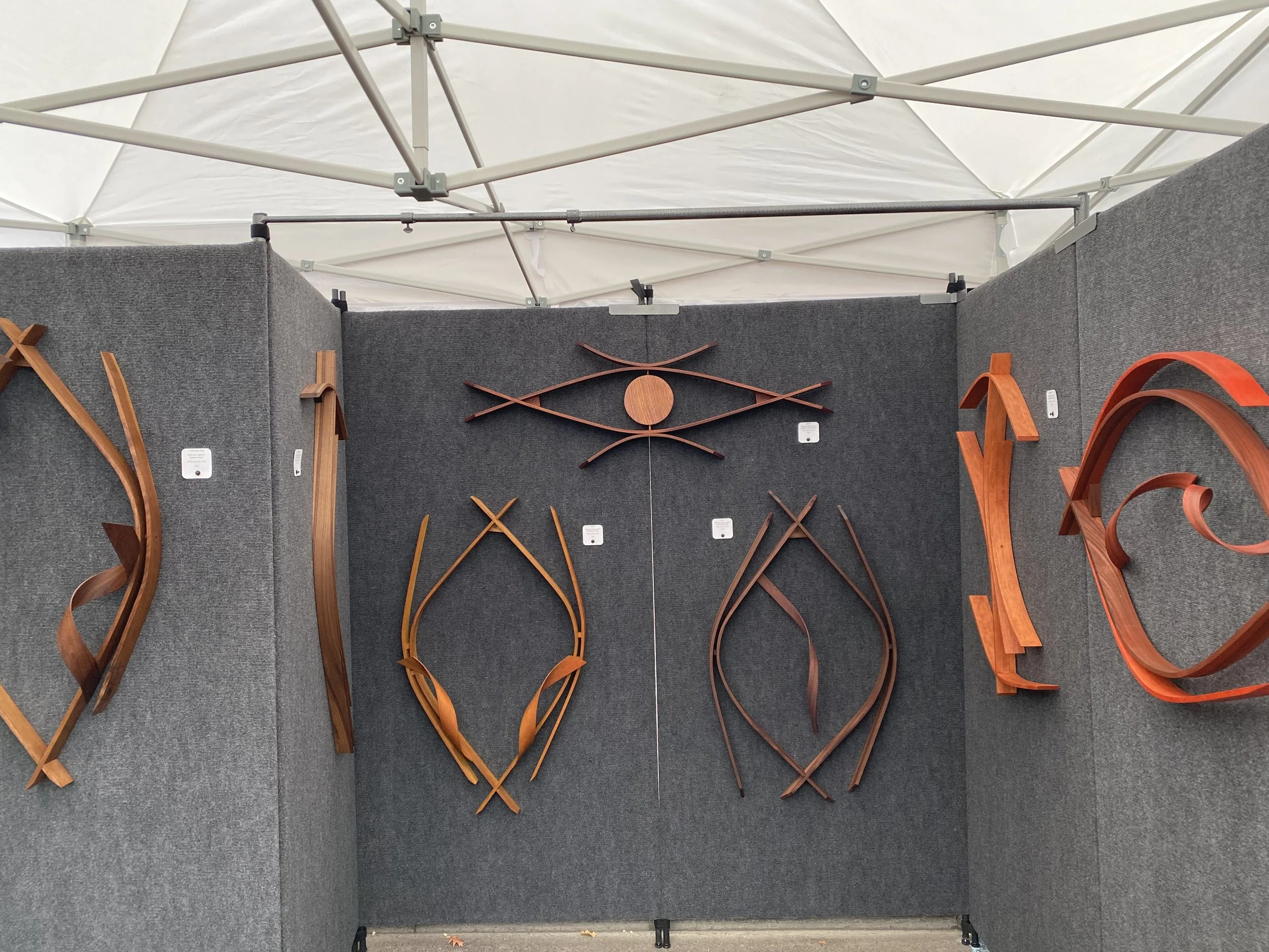 Display of various wooden sculptures mounted on a dark gray fabric-covered booth at an exhibition with a white tent overhead.