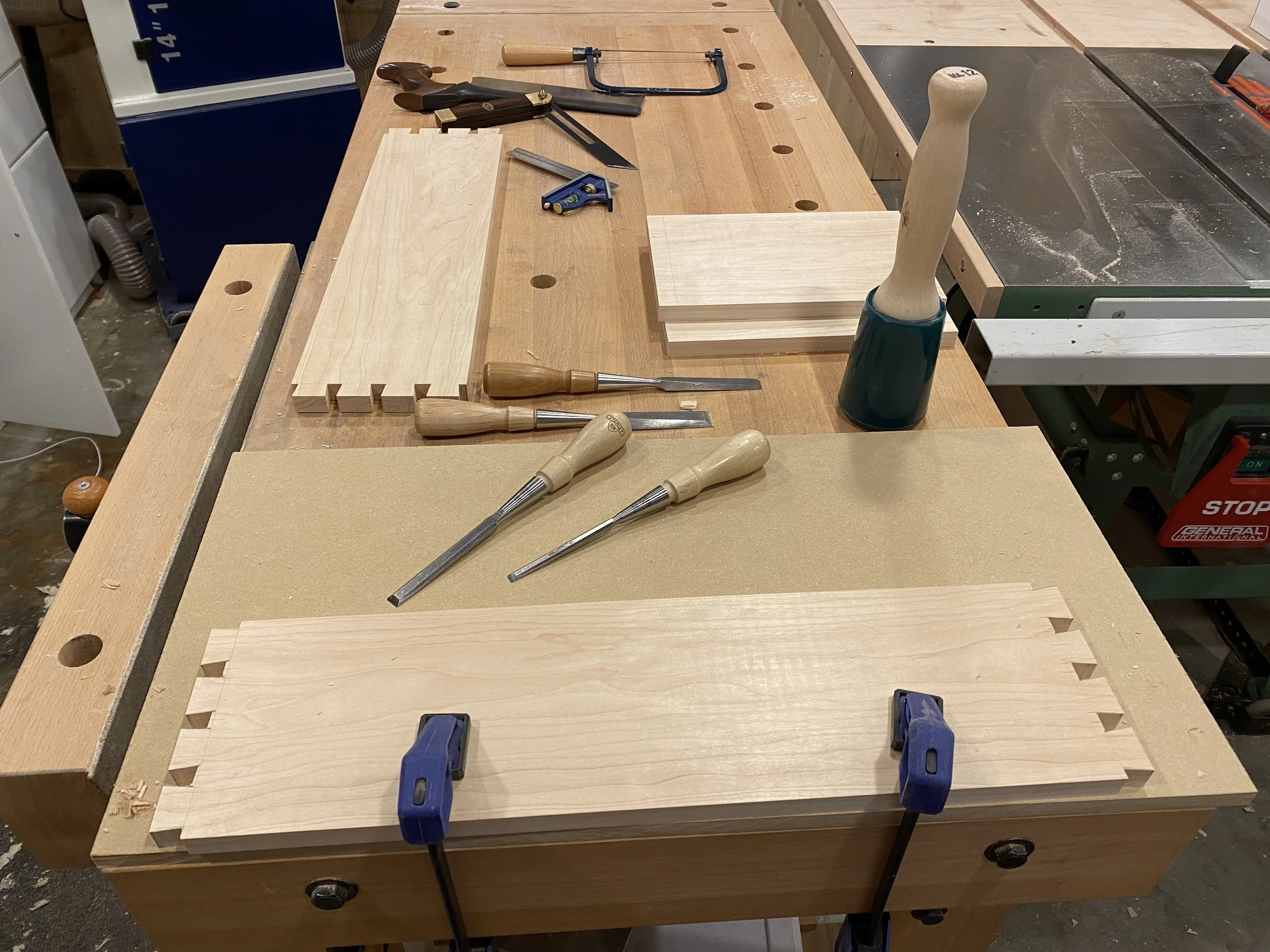 A woodworking workbench with various tools, including chisels, a hand saw, a square, clamps, and wood pieces, in a woodworking shop.