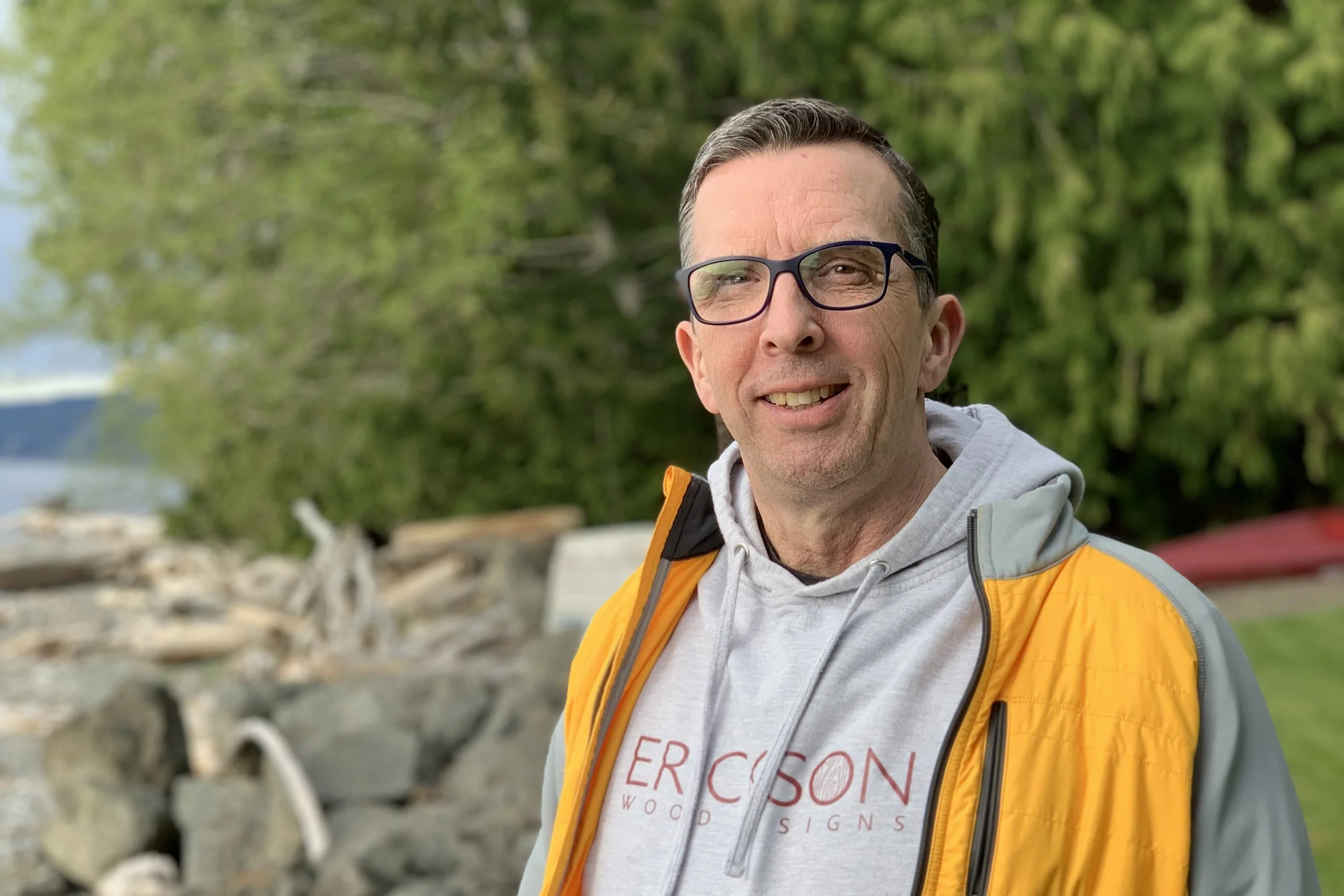 Smiling man with glasses and a yellow jacket outdoors near water and trees.