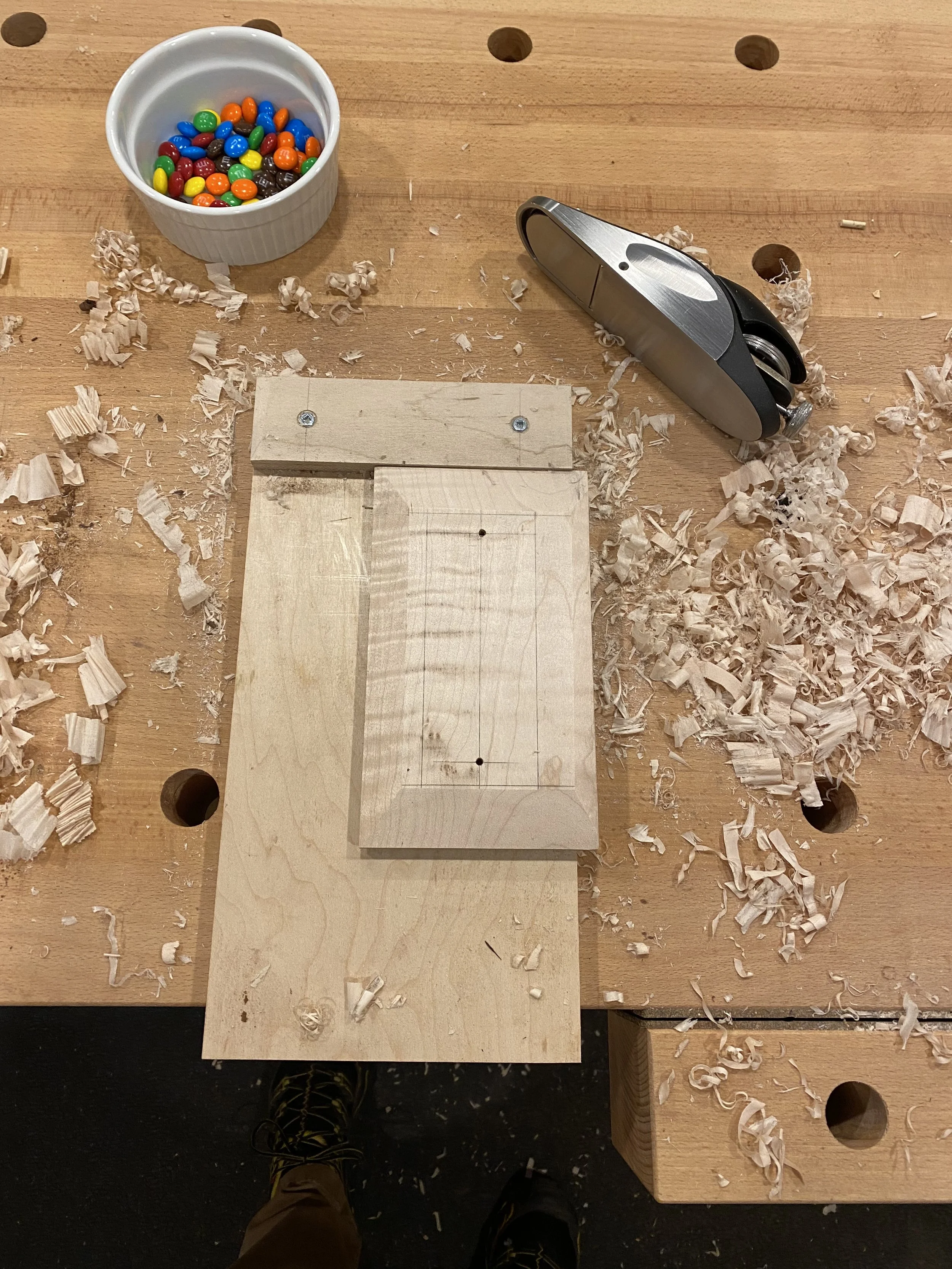 A wooden workbench with wood shavings scattered around, a small white bowl with colorful candy, and a metal nail clipper. The person's feet are visible at the bottom of the image.