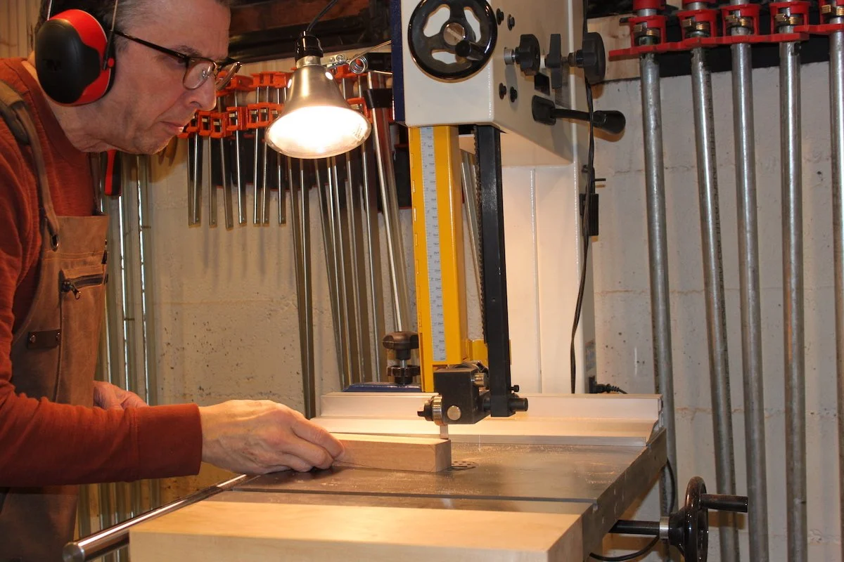 A man wearing safety glasses and ear protection is using a bandsaw to cut a piece of wood in a woodworking shop.