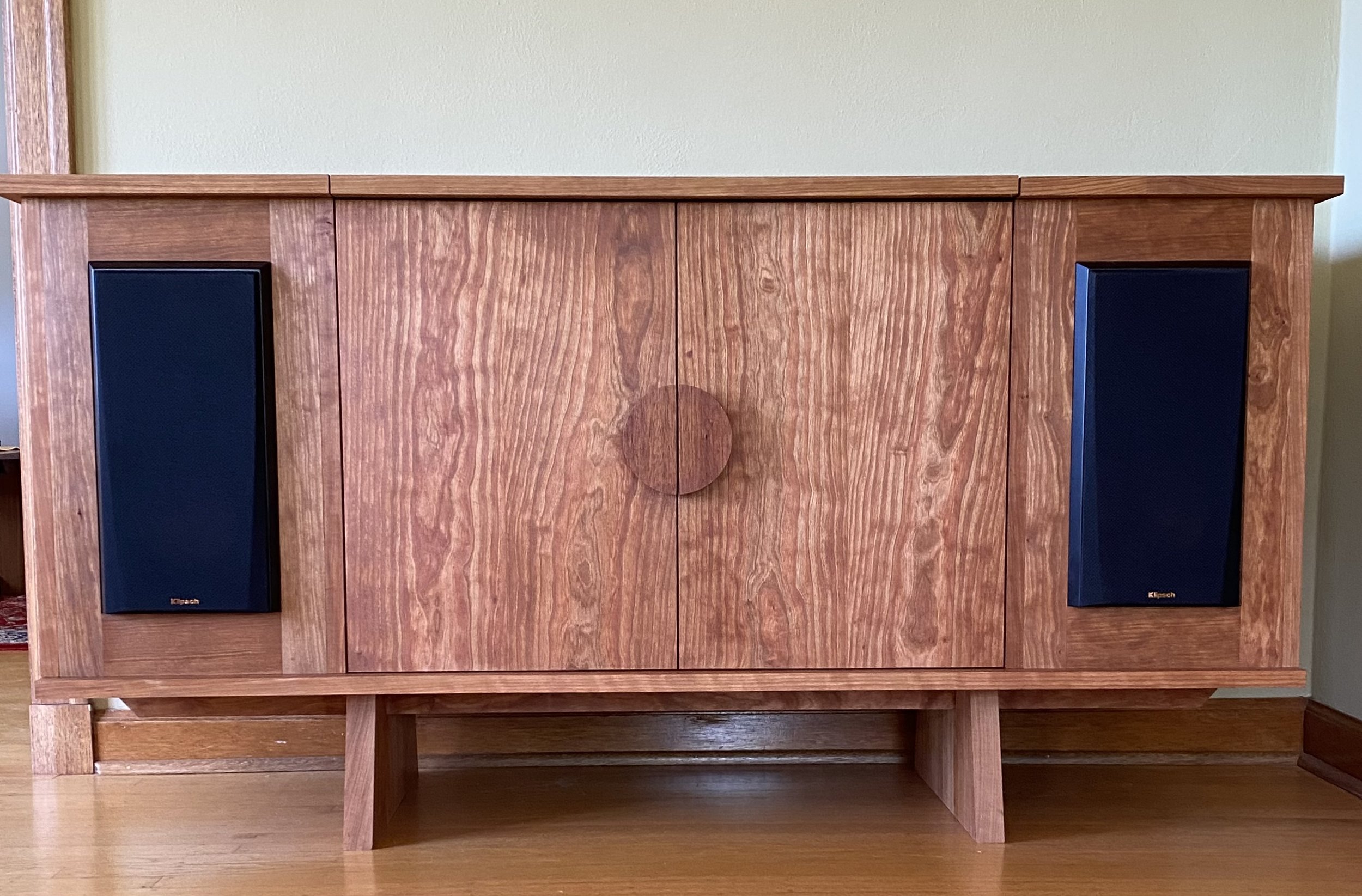 A wooden cabinet with two black Klipsch speakers on each side, sitting on a wooden floor with a pale green wall behind.
