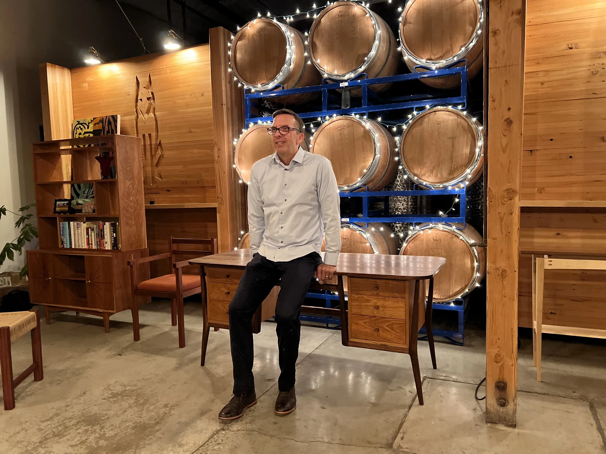 A man wearing glasses, a white dress shirt, black pants, and brown shoes is sitting on a table in a warmly lit room. Behind him, several hanging wine barrels are decorated with string lights, and the room features wooden walls and furniture.