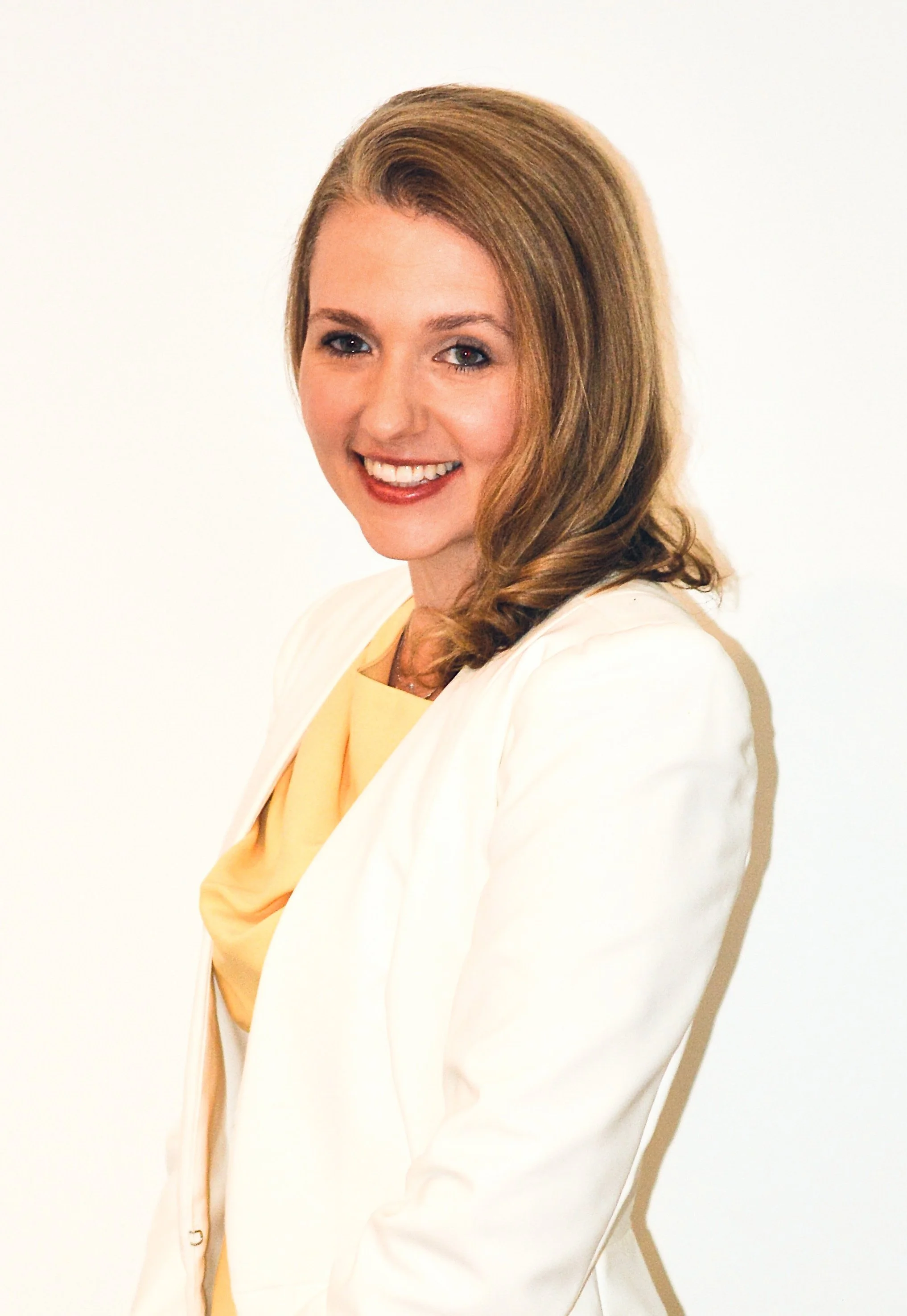 A woman with light brown, shoulder-length hair, smiling and wearing a light-colored blazer over a yellow top, standing against a plain white background.