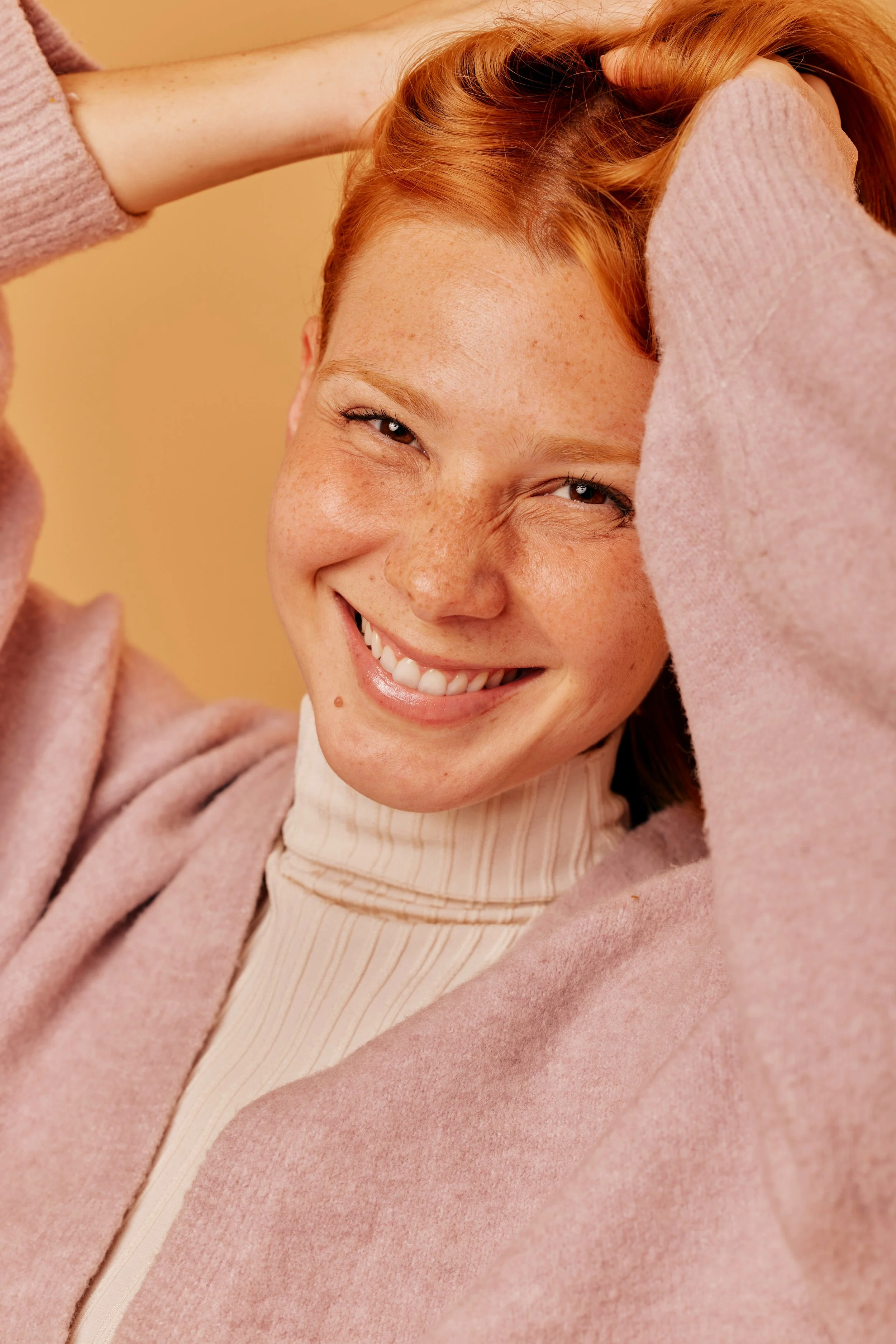Close-up of a smiling red-haired woman with freckles, wearing a beige turtleneck and pink sweater, with a yellow background.