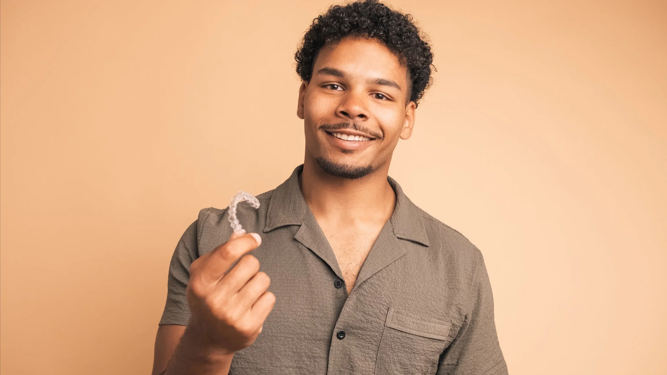 Young man with curly hair and a goatee smiling, holding a clear earplug in his hand, standing against a beige background.