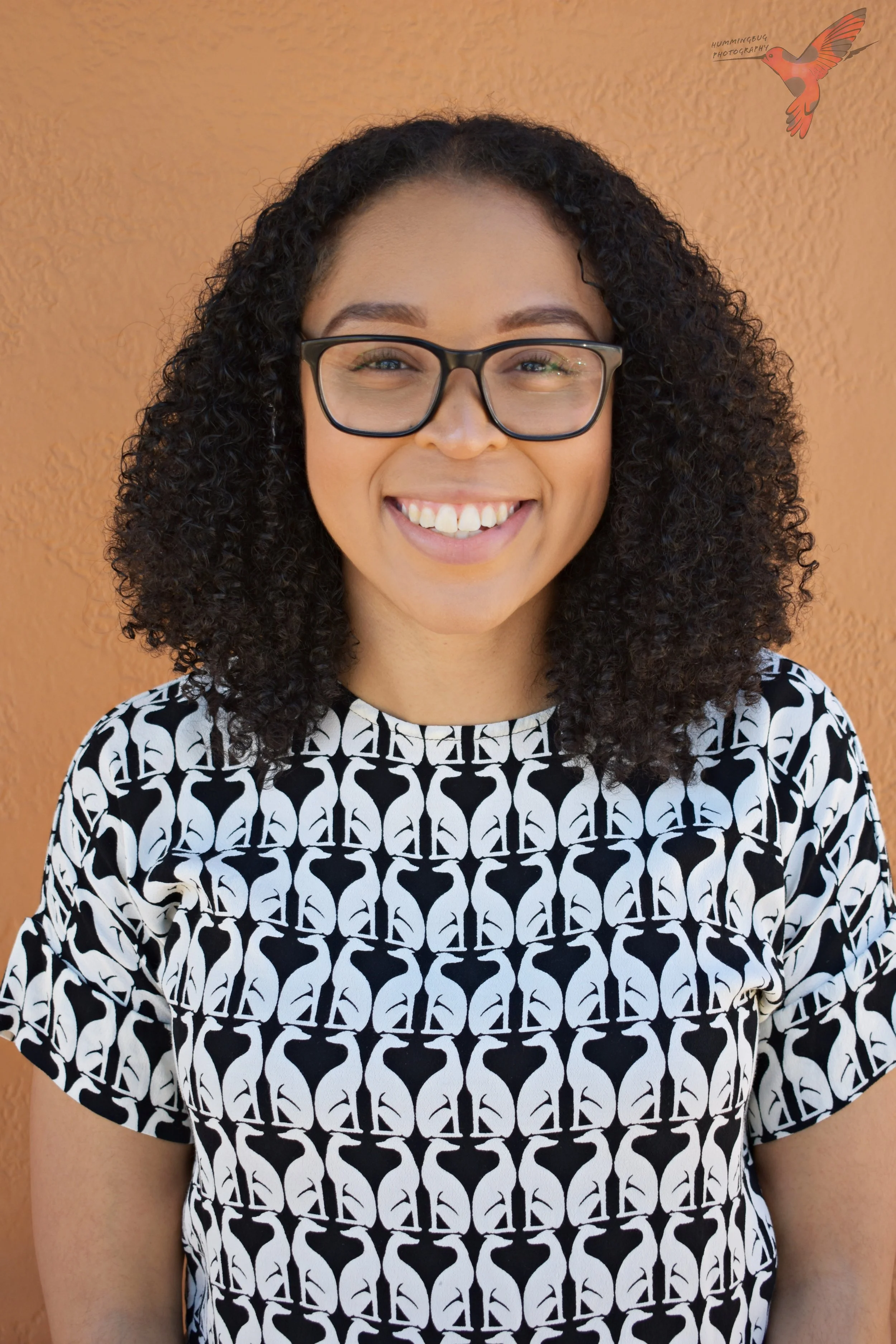 A woman with curly black hair, glasses, and a black-and-white patterned shirt smiling in front of an orange wall with a hummingbird sticker in the top right.