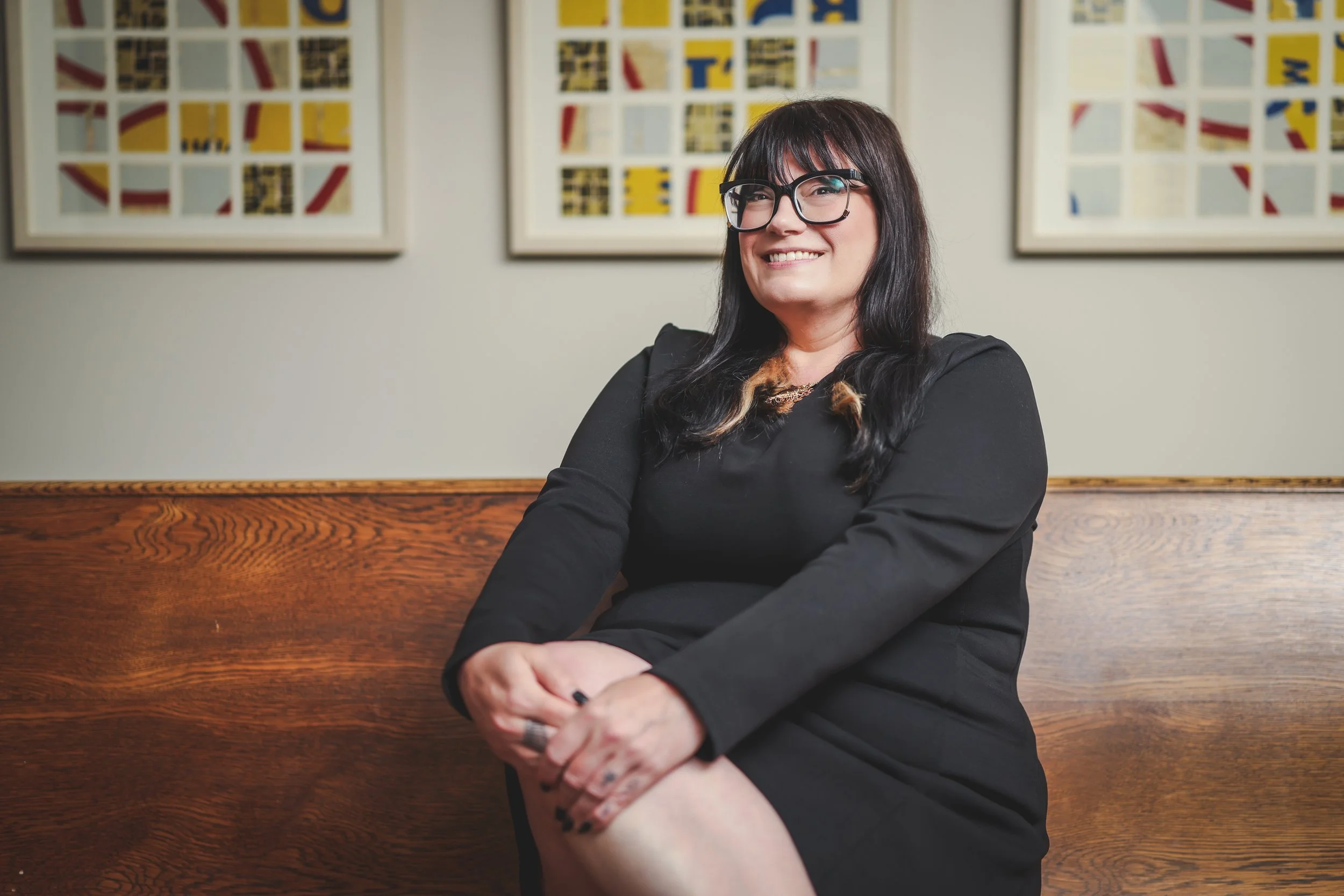 A woman with long dark hair, glasses, and a black top, smiling with crossed arms, sitting in front of abstract colorful framed artwork on the wall.