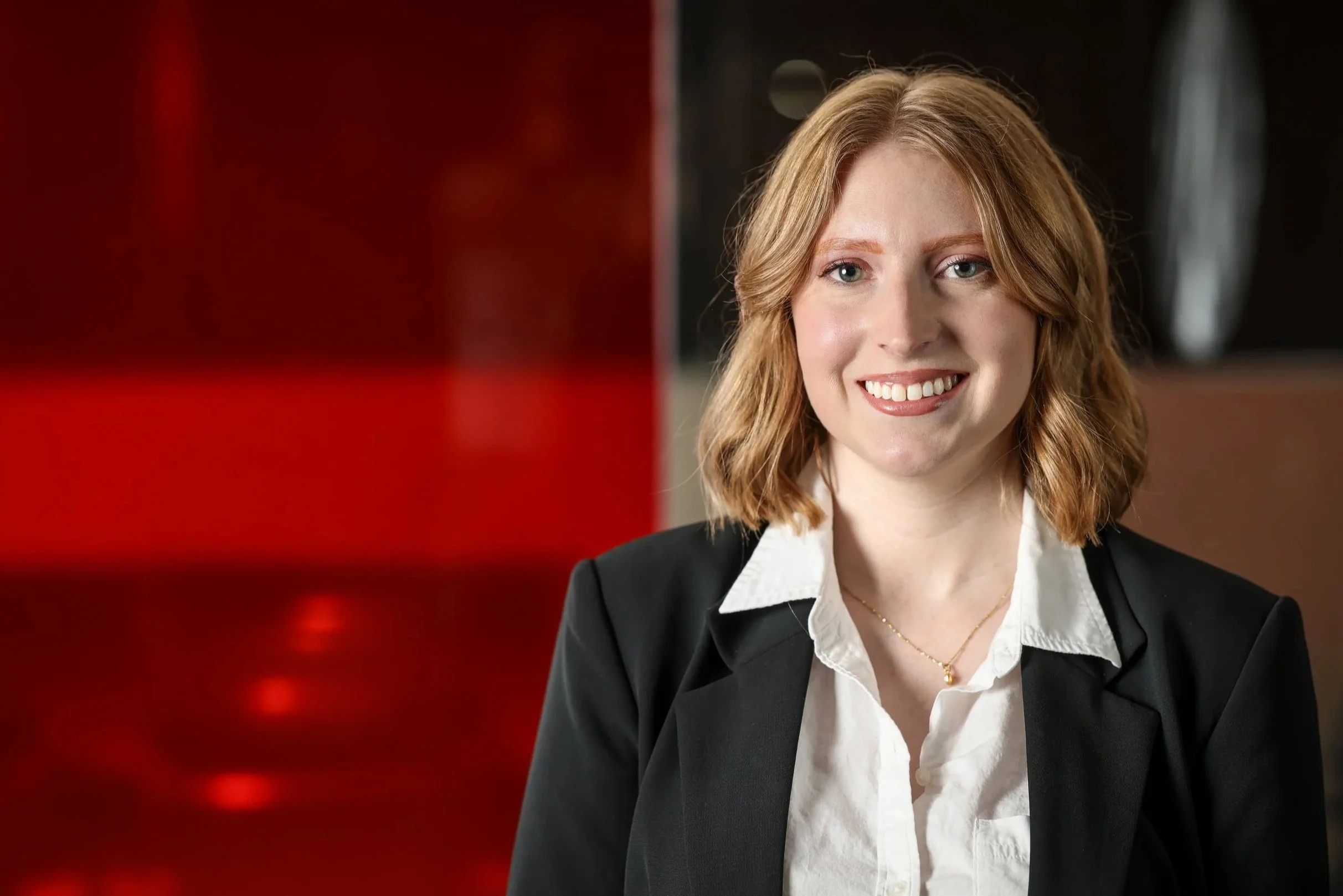 A woman with shoulder-length red hair wearing a black blazer and white shirt, smiling and looking at the camera, with a blurred background featuring red and black colors.