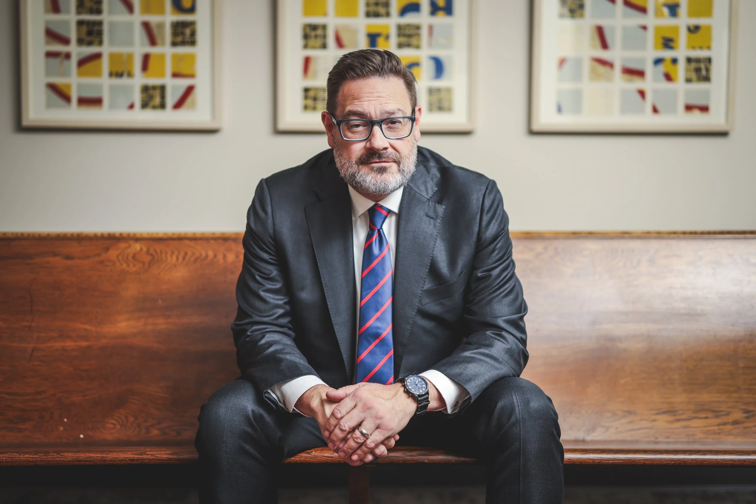 A man in a suit and tie sitting on a wooden bench with his hands clasped, wearing glasses and a wristwatch, in front of abstract artwork on the wall.