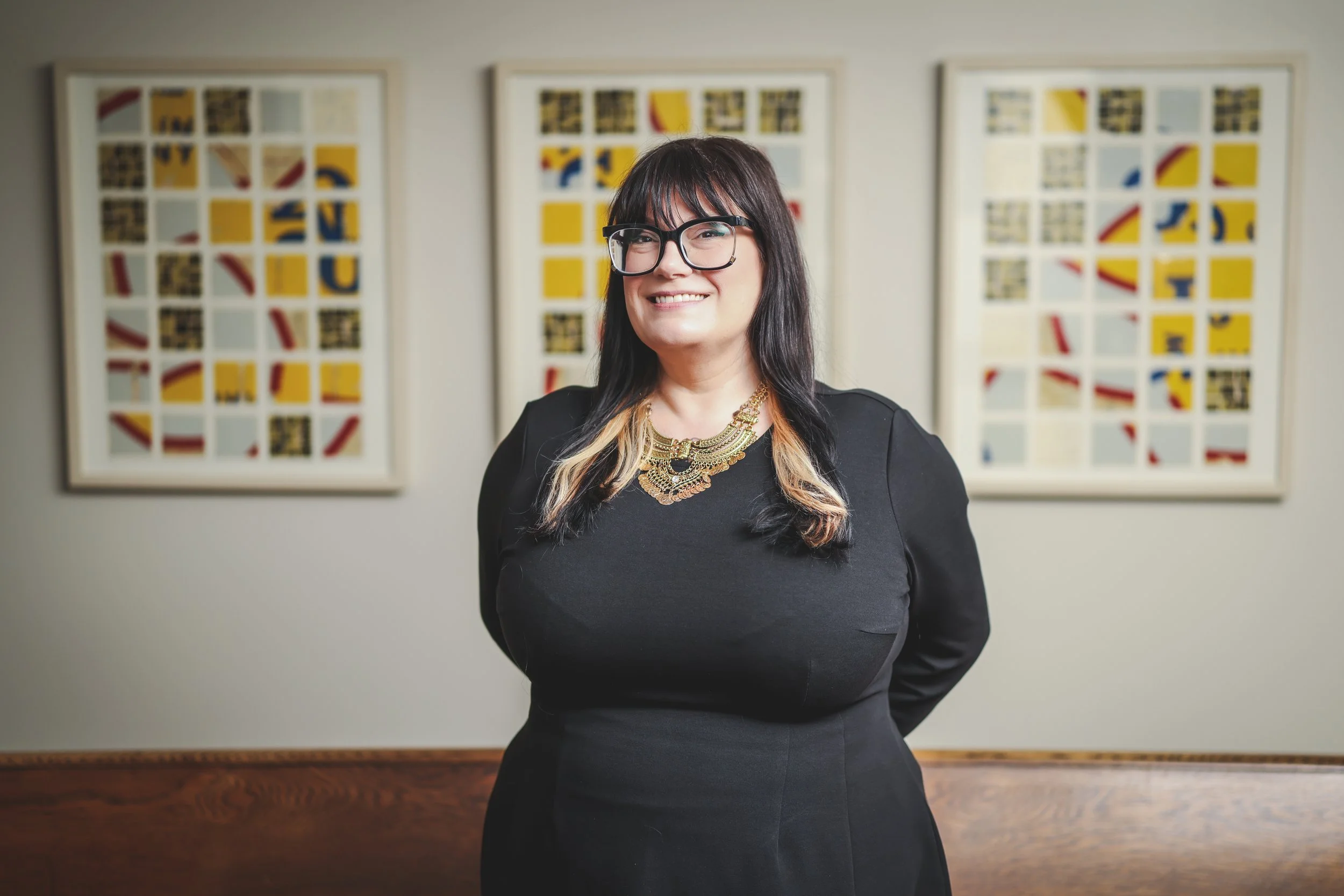 A woman with dark hair and glasses smiling, standing in front of three colorful abstract paintings on a gray wall.