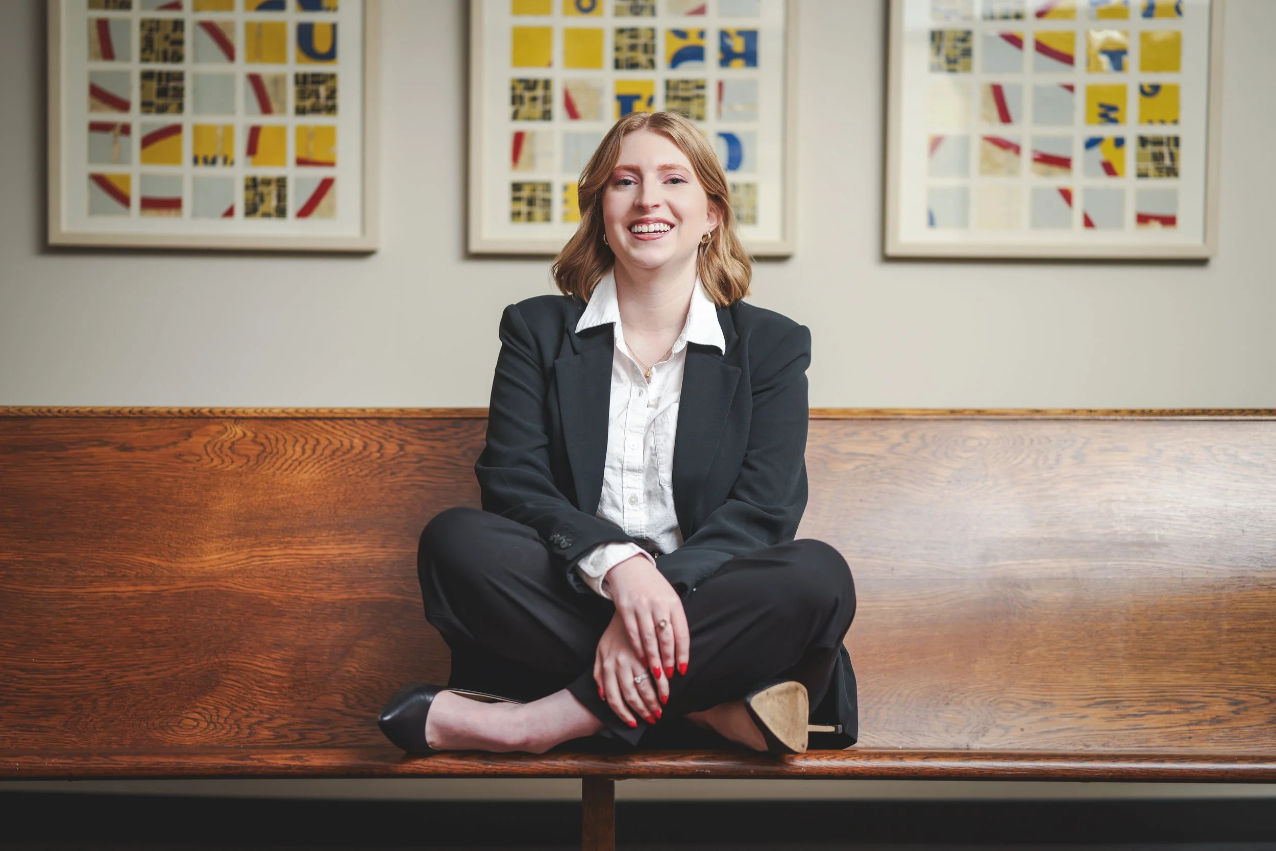 A woman with red hair, wearing a black blazer and white shirt, is sitting cross-legged on a wooden bench with a a smile. Behind her are three framed abstract art pieces with yellow, red, black, and blue colors on a gray wall.