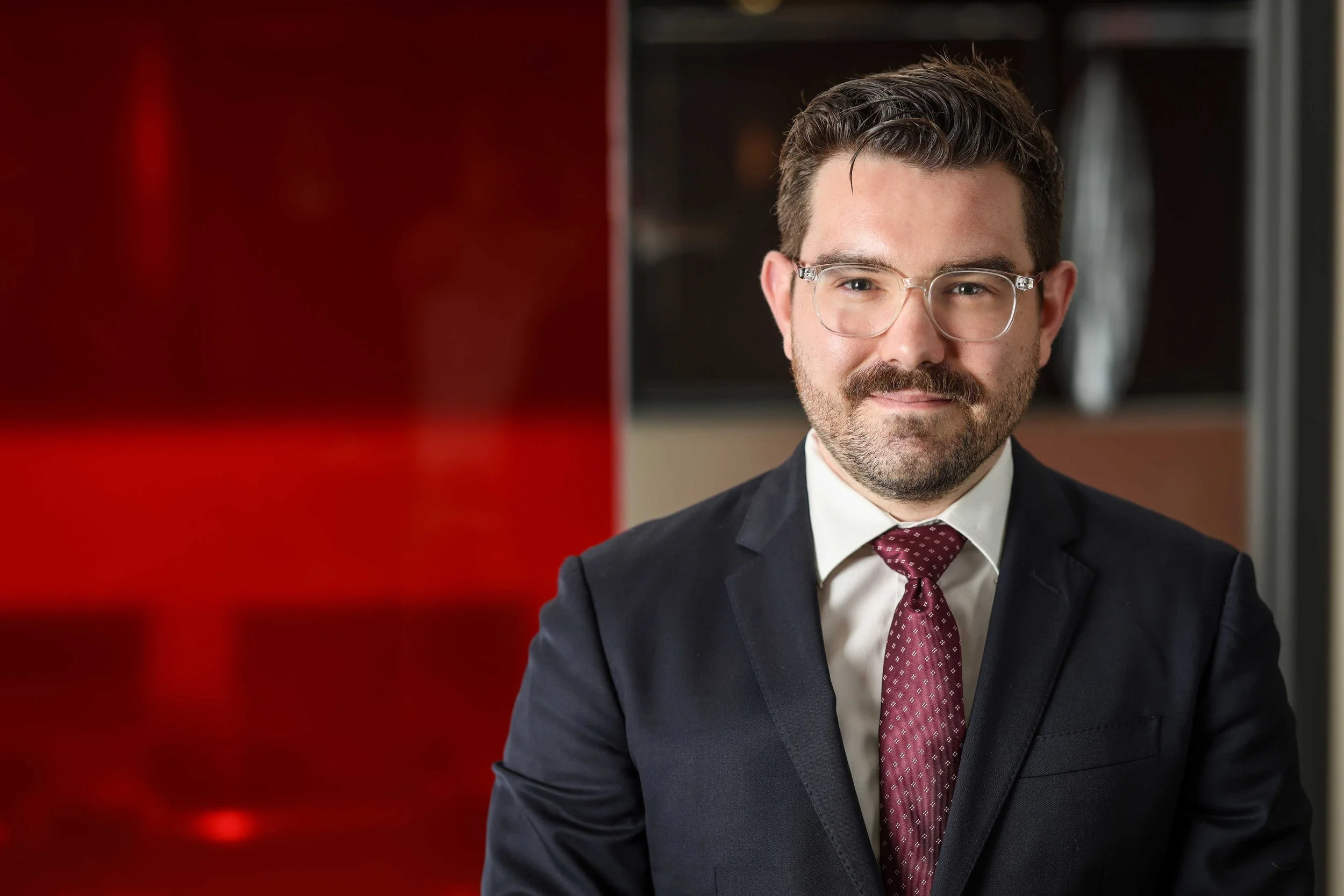 A man with glasses, dark hair, and a beard wearing a dark suit, white shirt, and maroon tie, sitting in front of a red and black background.