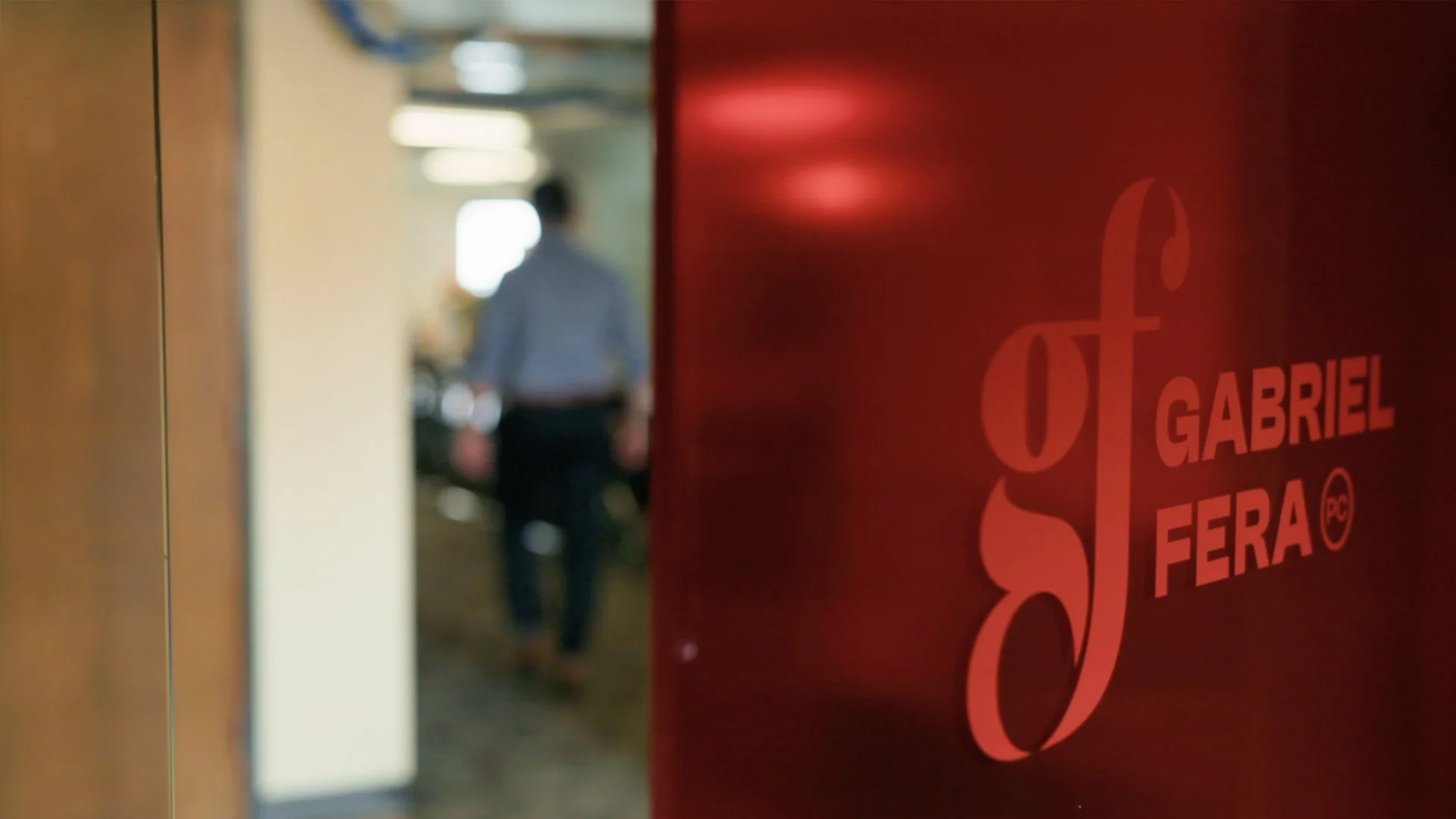 A red sign on a door reads 'St Gabriel FERA'. In the background, a person in a light blue shirt is walking in a hallway of an office or professional building, with blurred lighting and furniture.