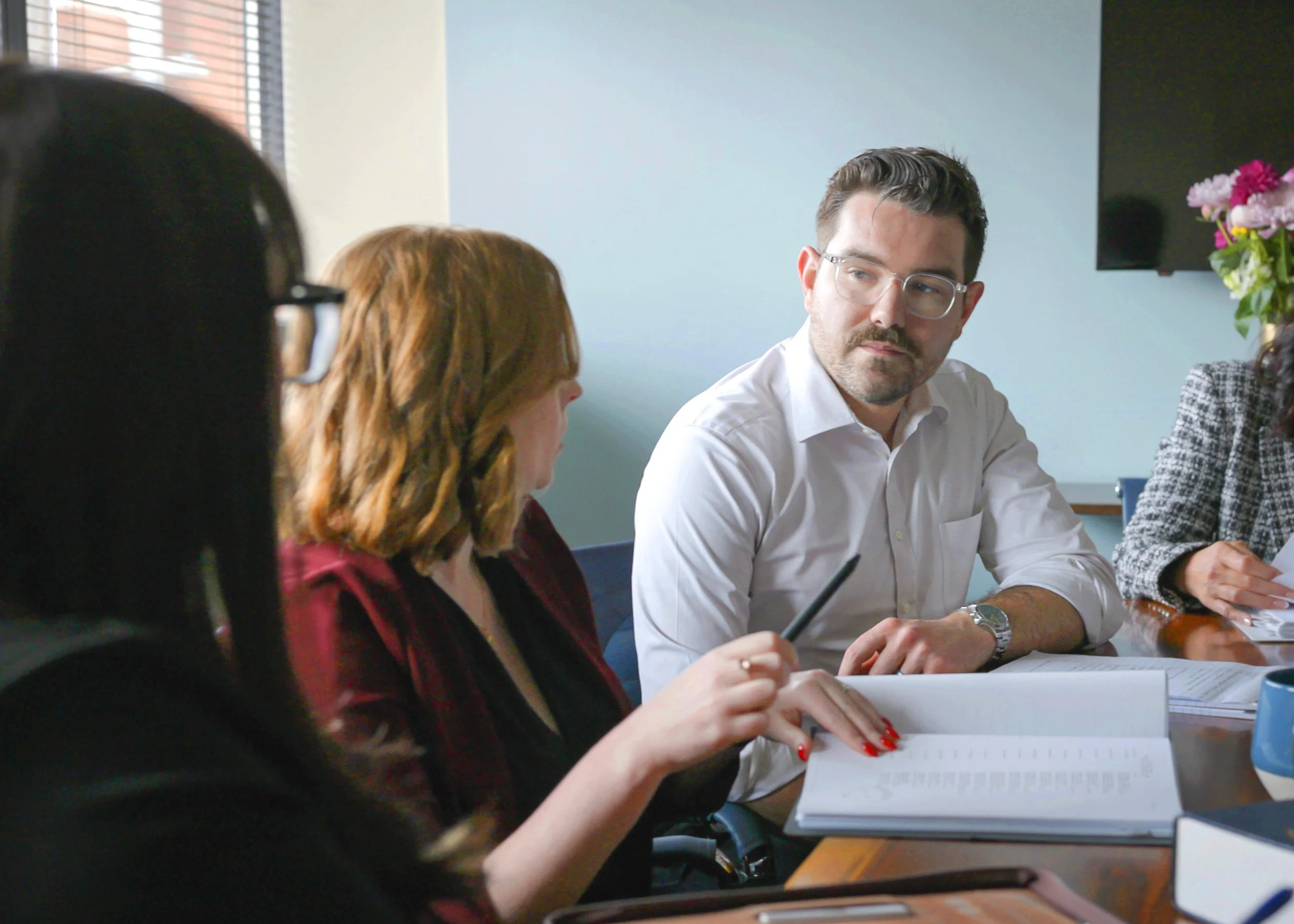 A group of people sitting around a table in a meeting room, engaged in discussion. One man in a white shirt and glasses looks to the side, while women with red and dark hair are taking notes and listening.