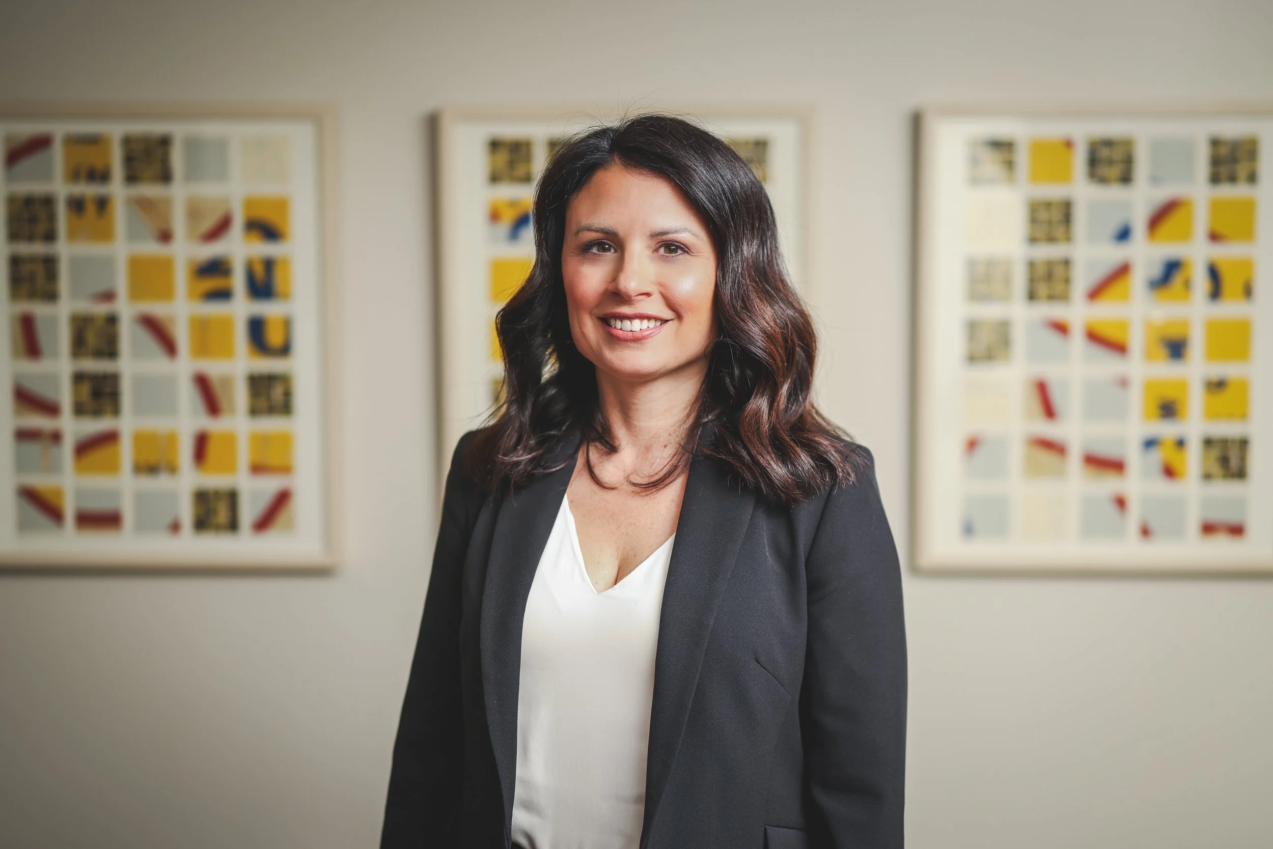 A smiling woman with dark, wavy hair wearing a black blazer and white top, standing in front of a white wall with three colorful abstract art pieces.