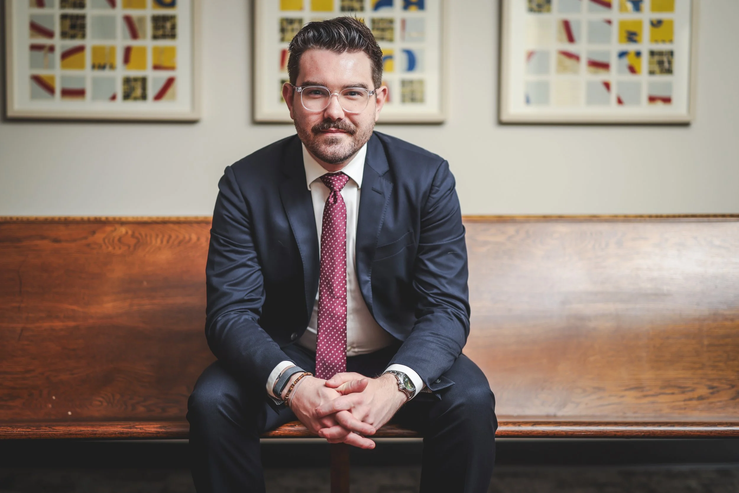 A man in a navy suit and maroon dotted tie sitting on a wooden bench in front of framed geometric art on a light-colored wall.