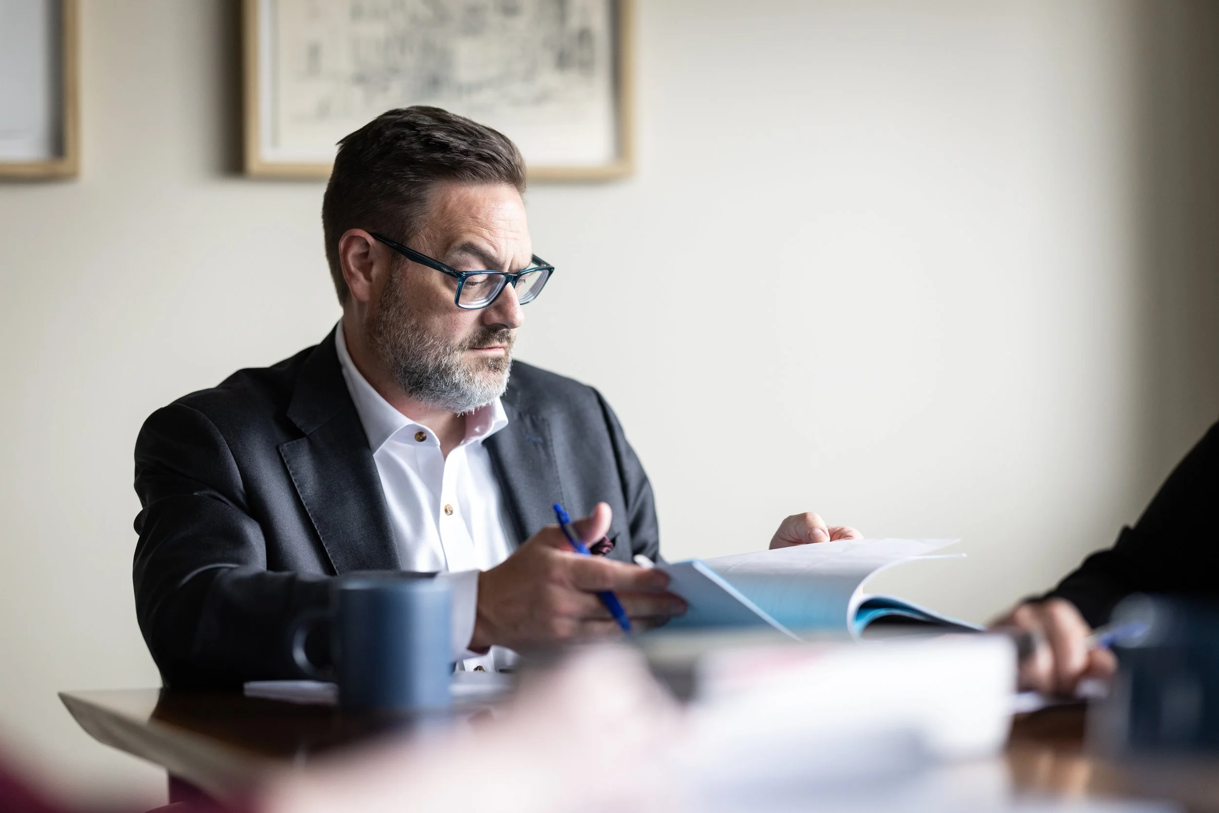 A man with dark hair, glasses, and a beard, wearing a suit and white shirt, is sitting at a table looking at papers, holding a blue pen.