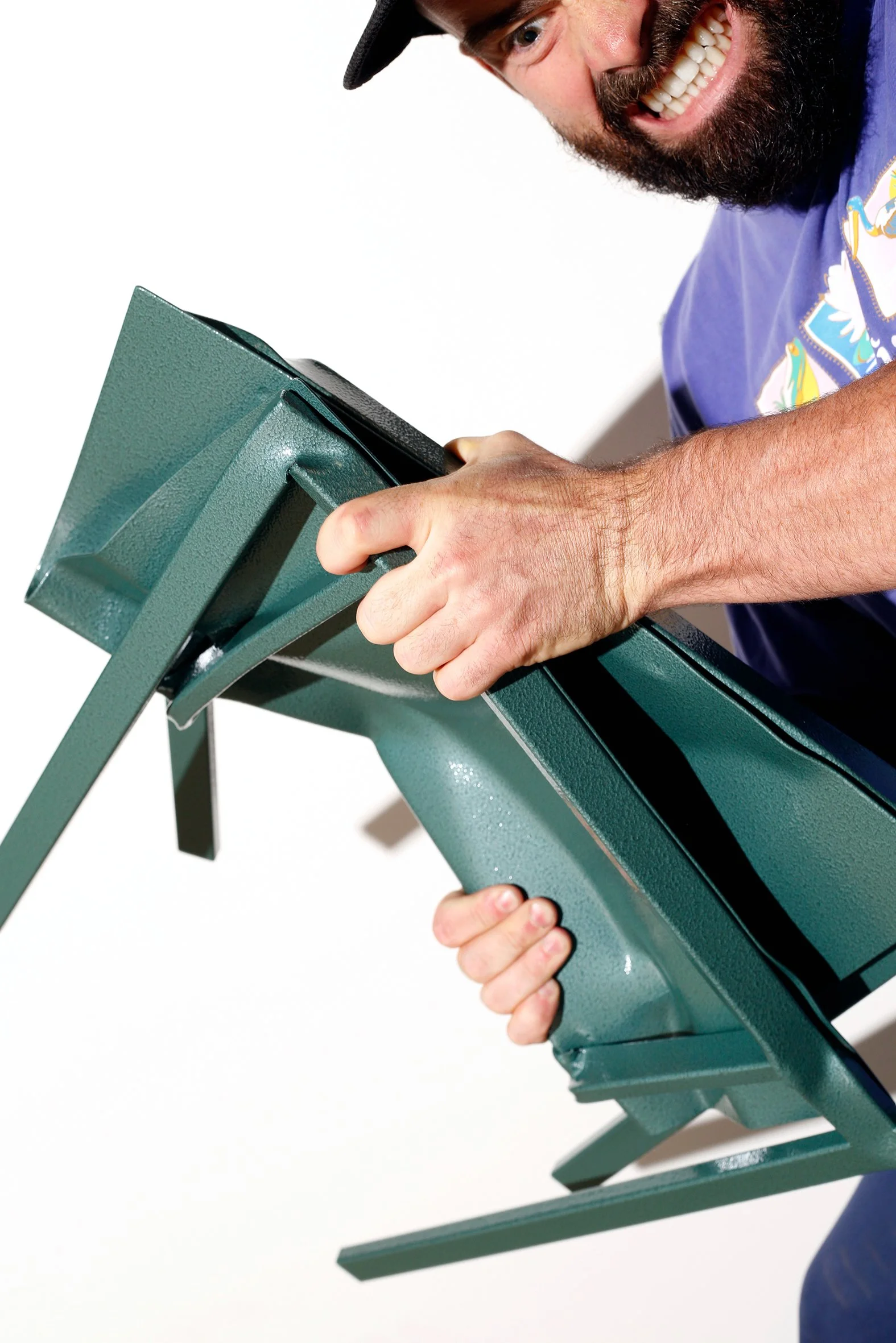 A man with a beard, wearing a colorful jacket and a black cap, is smiling while holding a green metal chair or bench.
