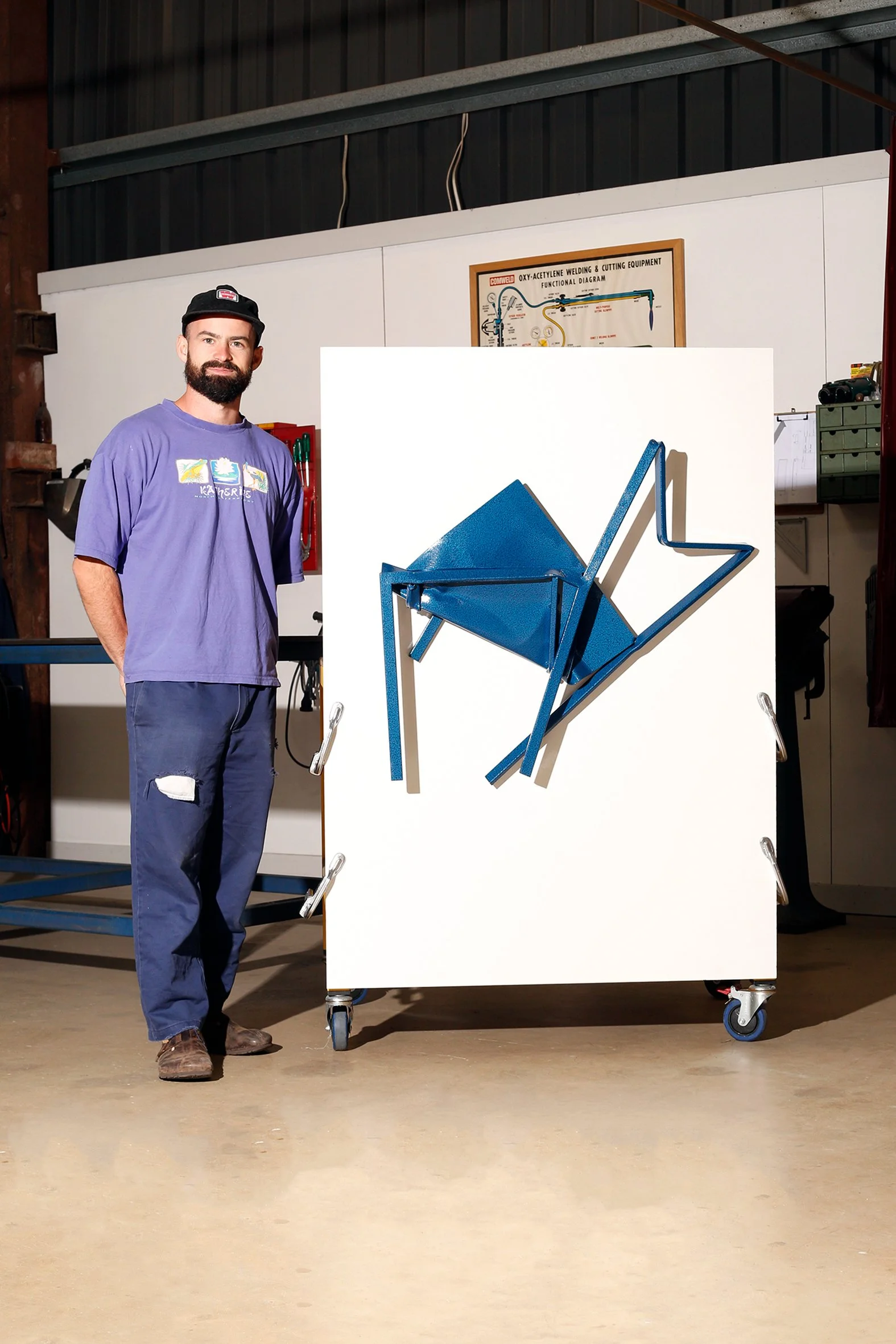 A man with a beard and a black beanie standing next to an art installation of a blue geometric chair mounted on a white background. The man is wearing a purple T-shirt and ripped blue jeans, and is indoors in a workshop or studio setting.