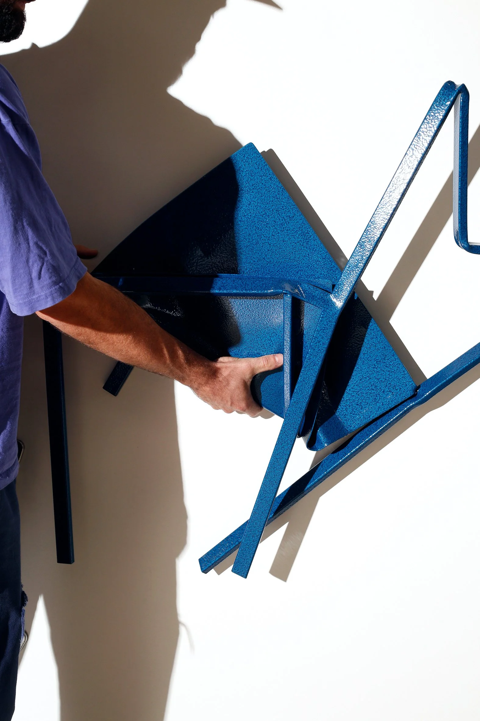 A person wearing a purple shirt holds a stack of blue metal chairs against a plain white background. Shadows are cast on the wall from the chairs and person.