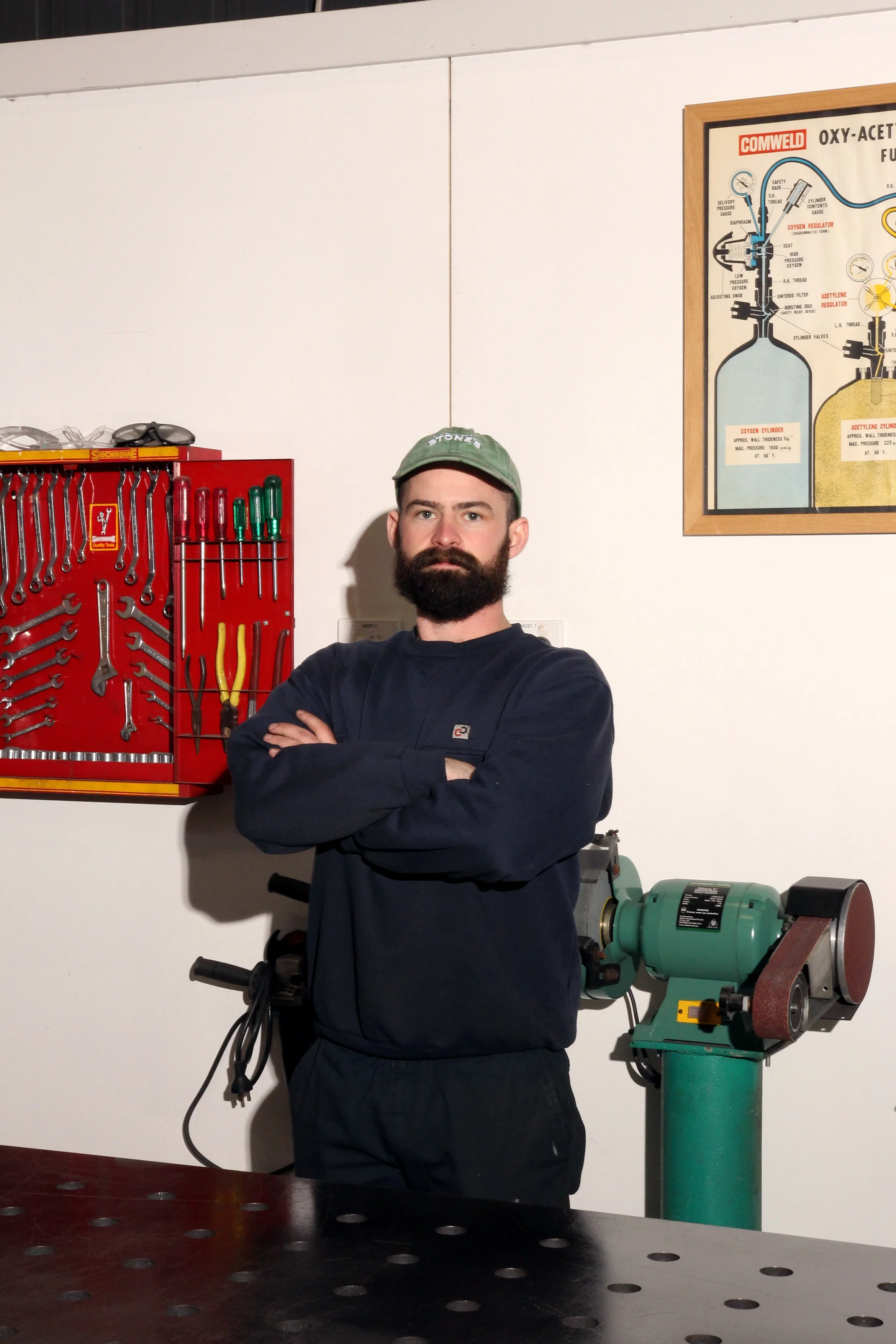 A man with a beard wearing a green cap and dark clothing standing with crossed arms in a workshop or garage, surrounded by tools and equipment, with a diagram of oxygen and acetylene cylinders on the wall.