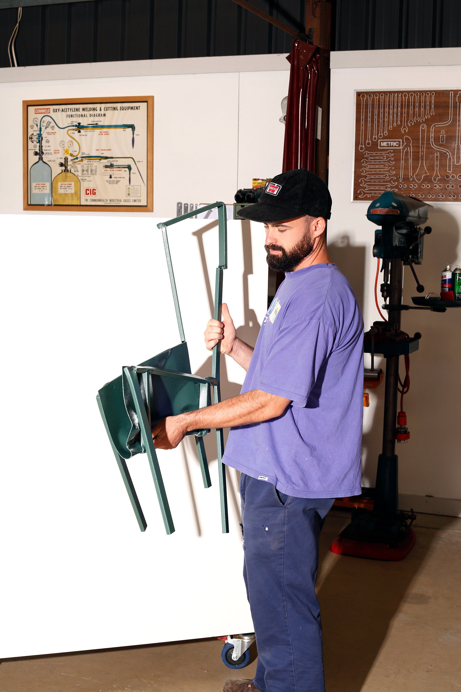 A man working in a workshop with tools, standing next to a white partition and holding a green metal frame. Behind him are posters, one showing welding equipment and another with a wrench diagram, along with machinery and tools on shelves.