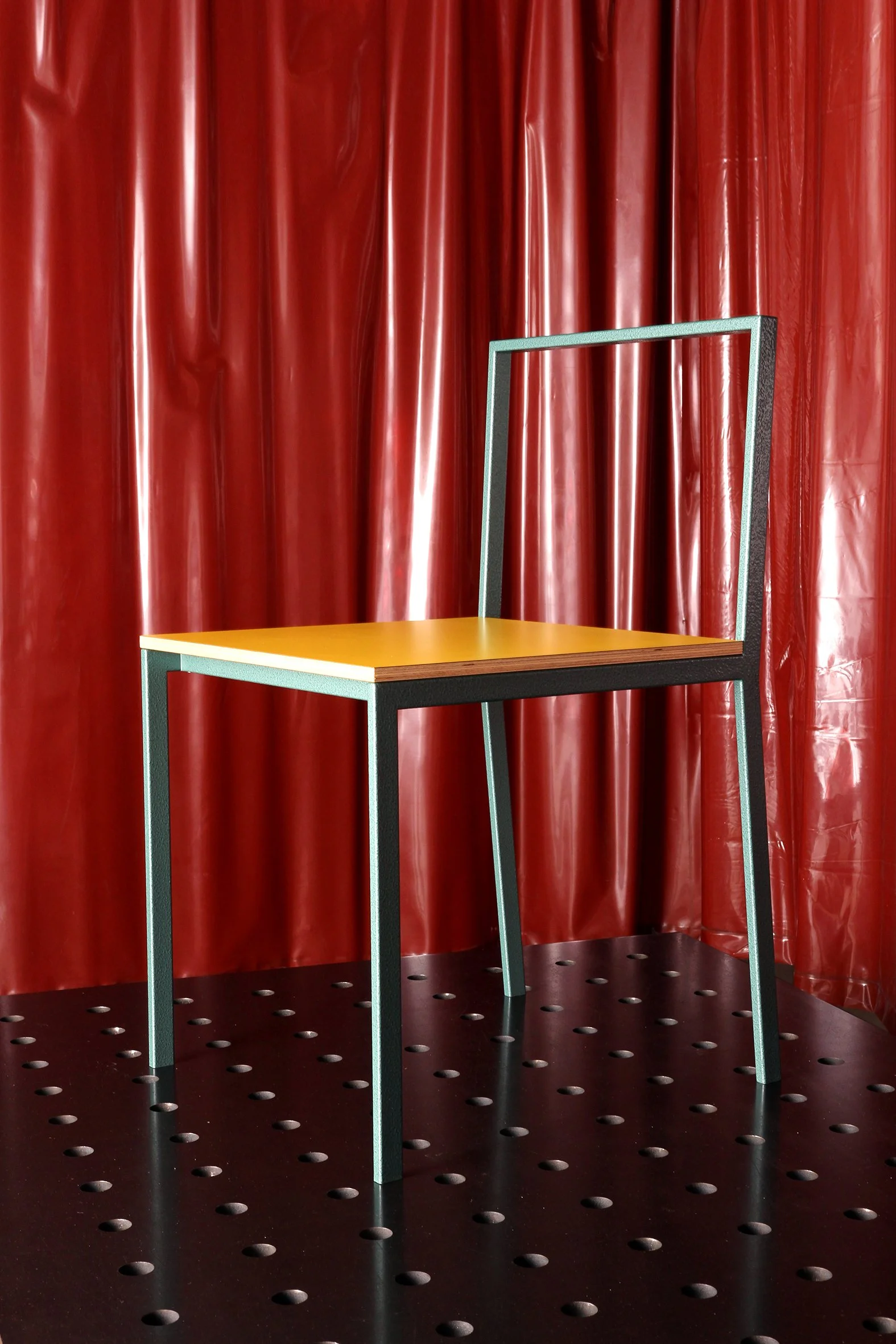 A mustard yellow chair with a black metal frame, placed in front of a red curtain, on a black perforated floor.