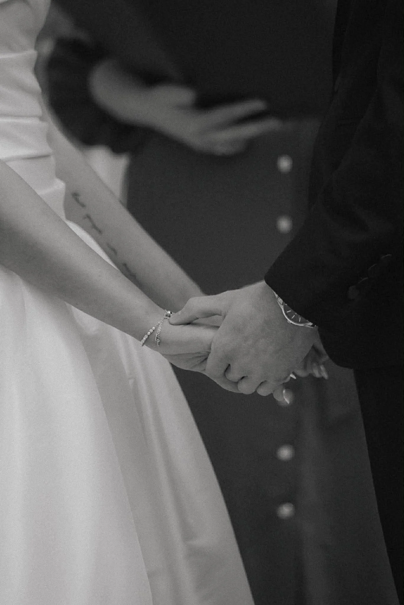 Close-up of two people holding hands, one wearing a bracelet, with a white surface below and a dark blurred background.