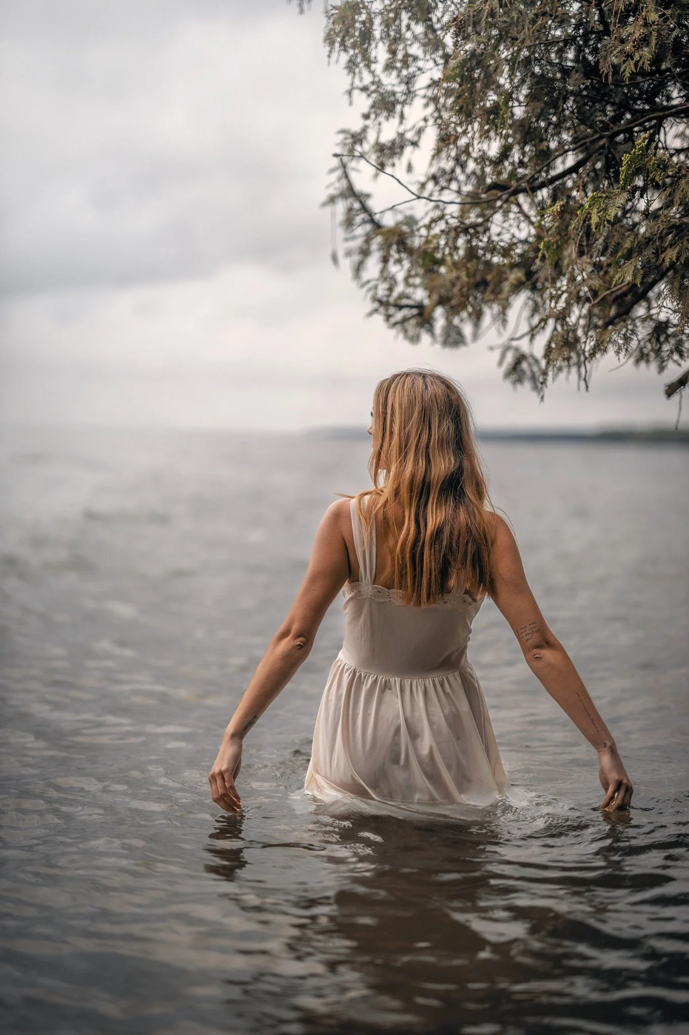 A woman with long, wavy, blonde hair standing in shallow water at the beach, wearing a white dress with her arms outstretched. Overhanging tree branches are visible in the upper right corner. The sky is overcast, creating a calm, serene atmosphere.