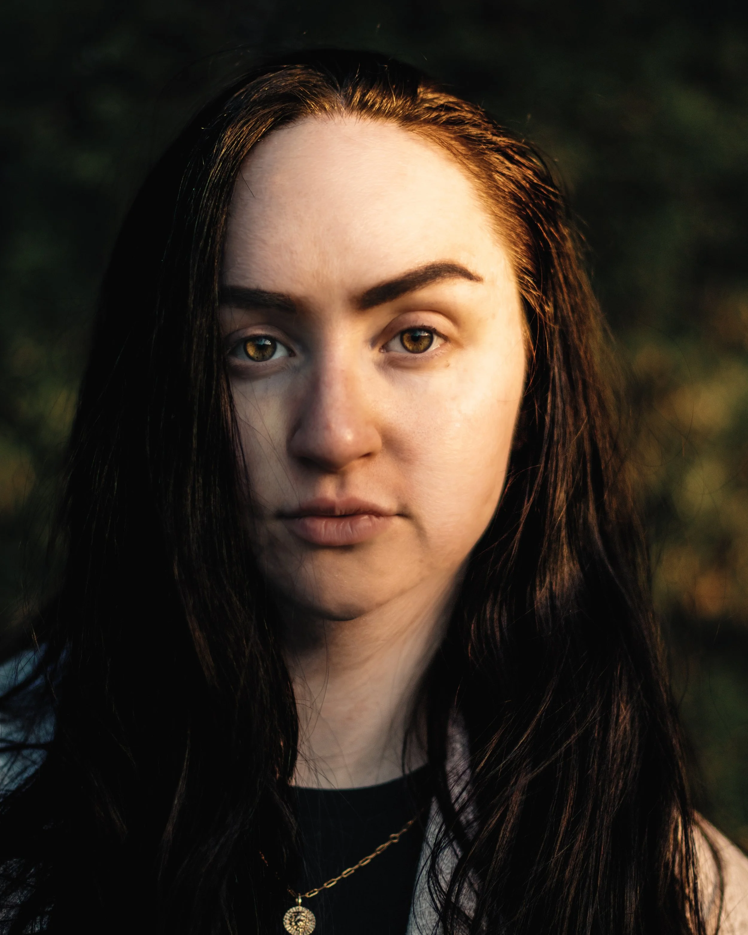 Close-up of a woman's face with striking hazel eyes, dark hair, and a gold necklace with a pendant, outdoors during golden hour.
