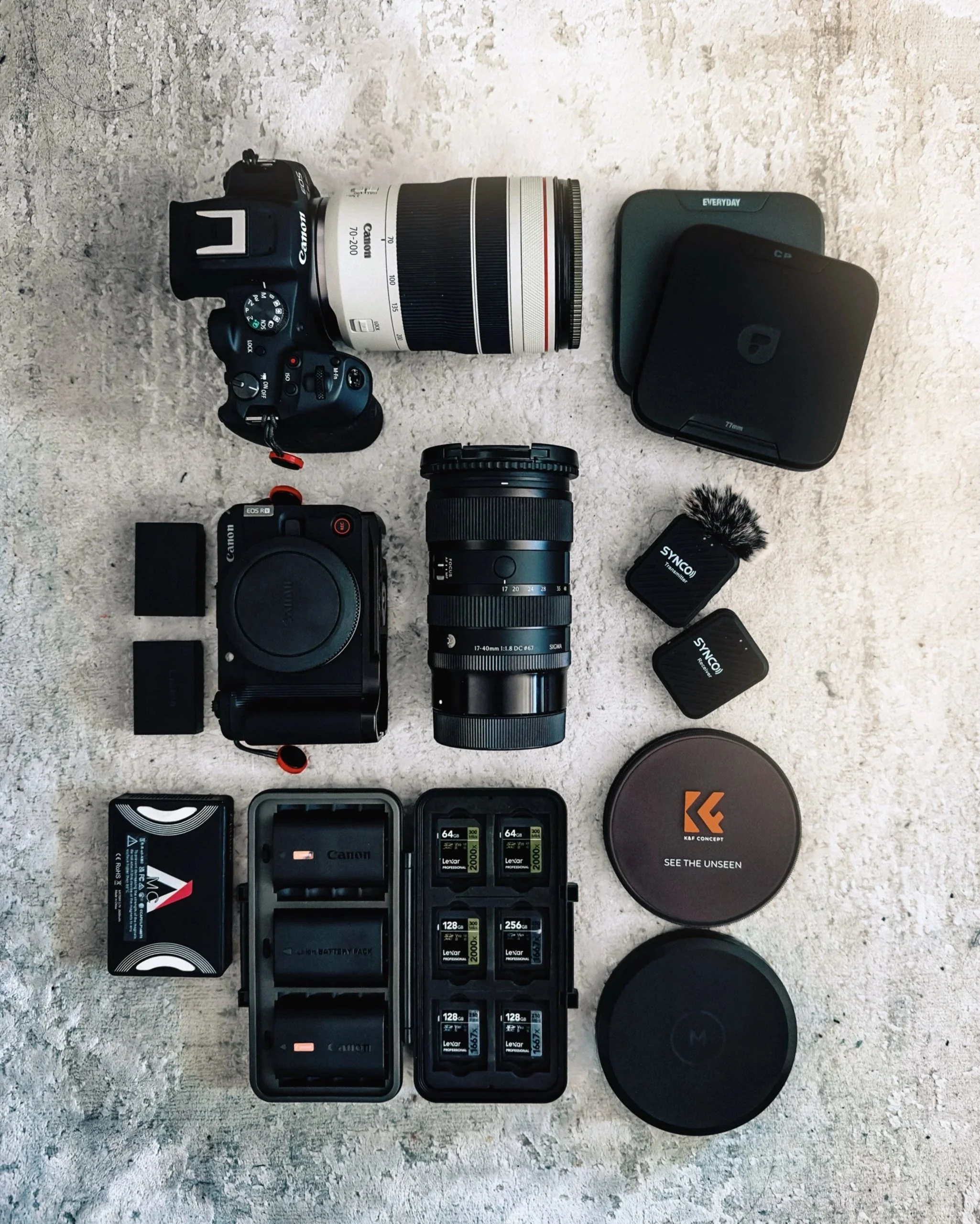 A collection of camera equipment and accessories laid out on a textured surface, including a Canon camera with a white telephoto lens, a second black camera body, two camera lenses, a camera battery pack, two SD memory cards, a microphone, two extern