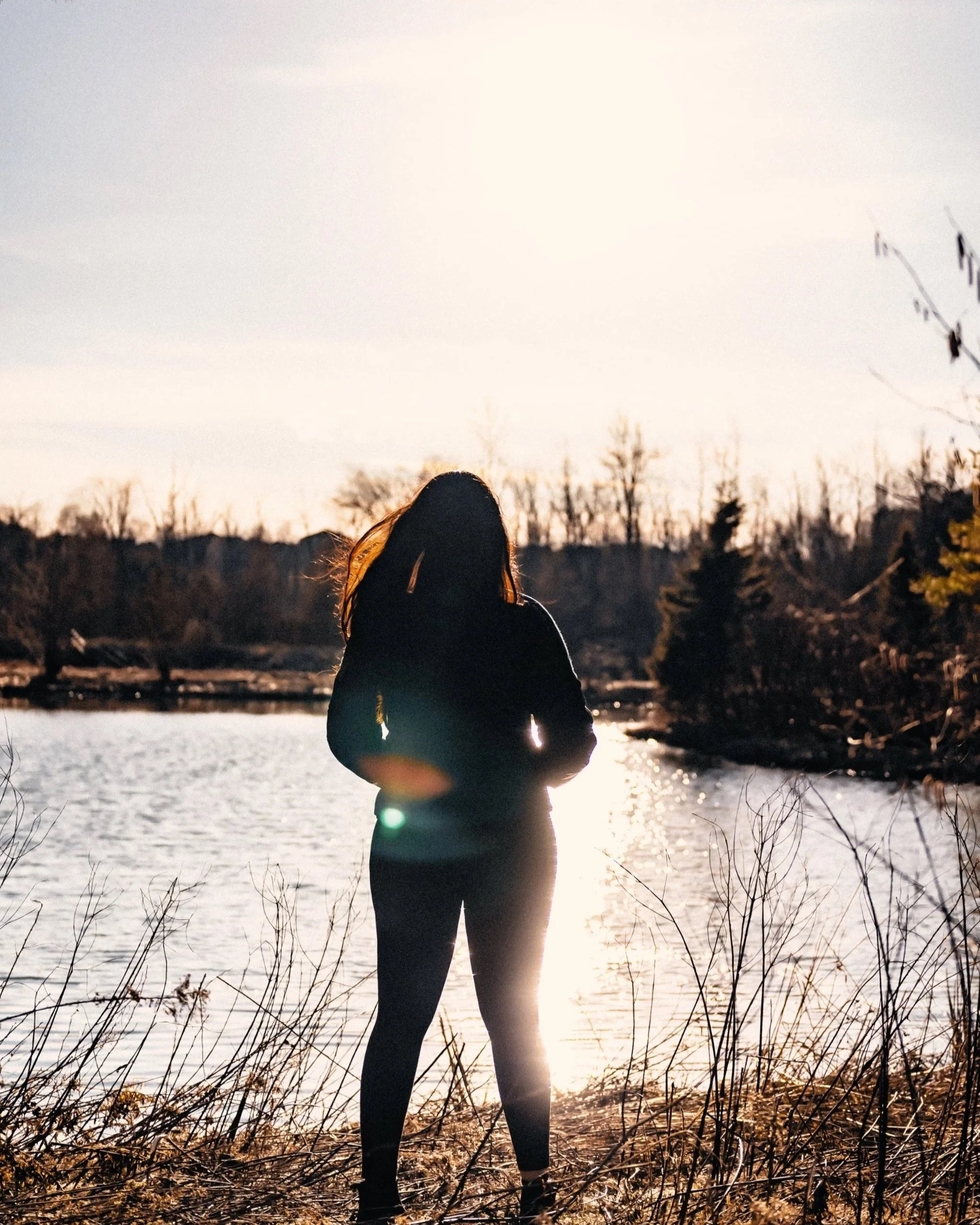Person standing near a body of water during sunset, with tall grass and trees in the background.