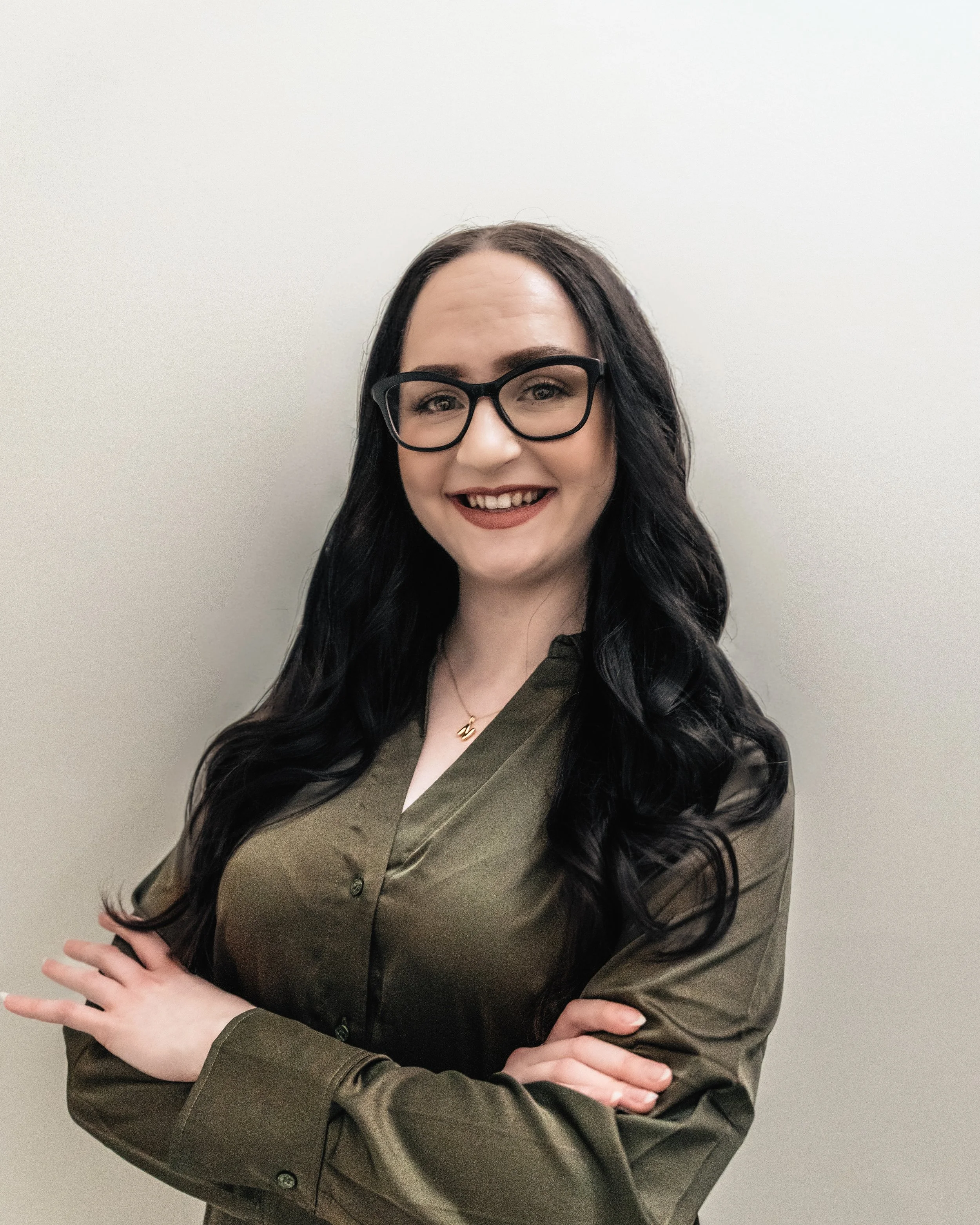 A woman with long dark hair, glasses, and a bright smile, standing against a plain white wall, crossing her arms.