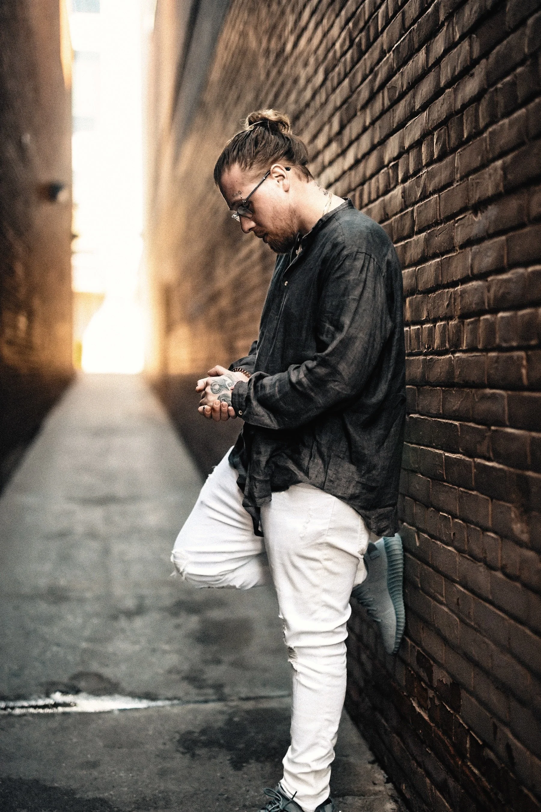 A young man with glasses, a top knot hairstyle, and tattoos on his hands, standing against a brick wall in an alleyway, dressed in a black shirt and white ripped jeans.