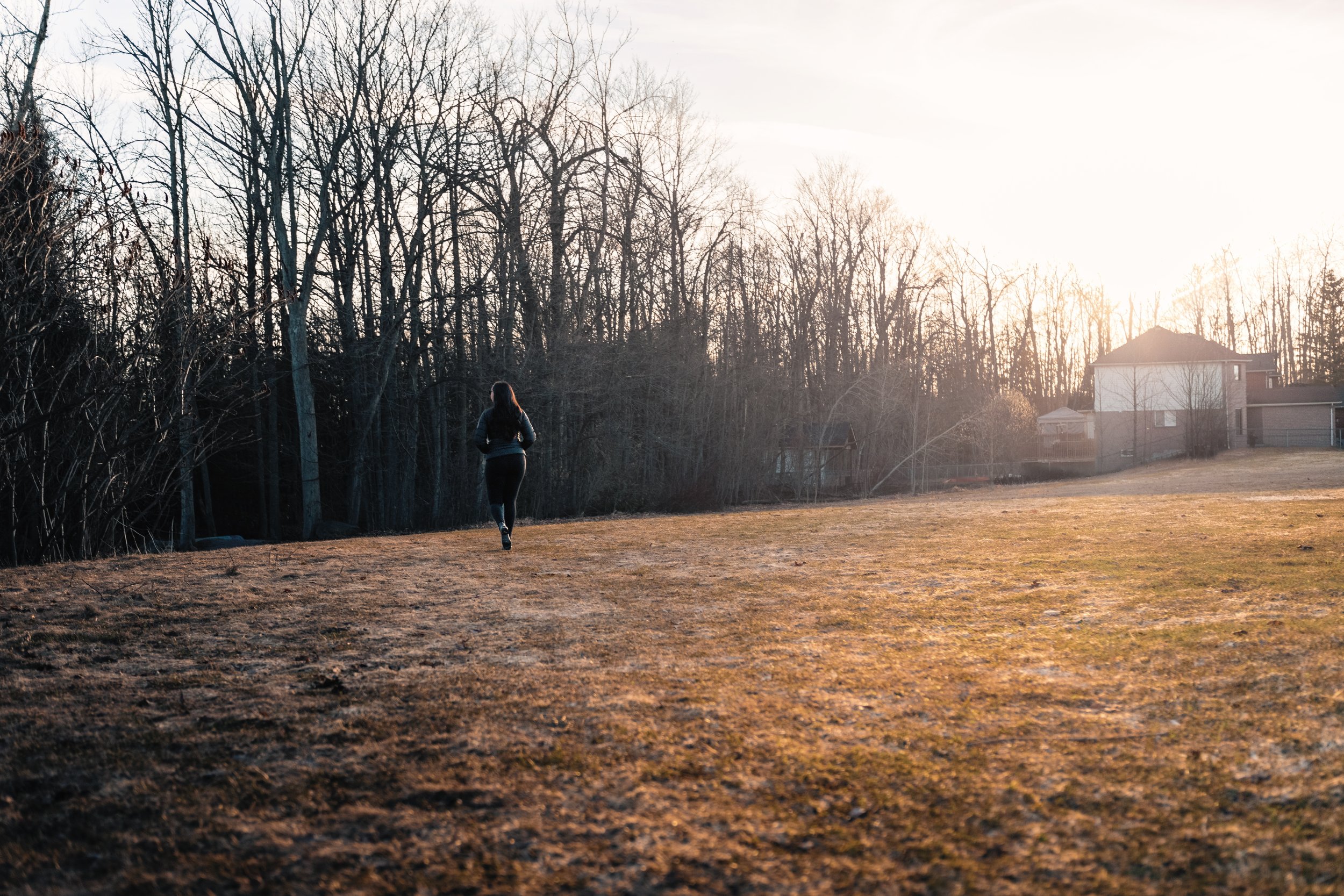 A person jogging in a park near houses with leafless trees in the background during sunset or sunrise.