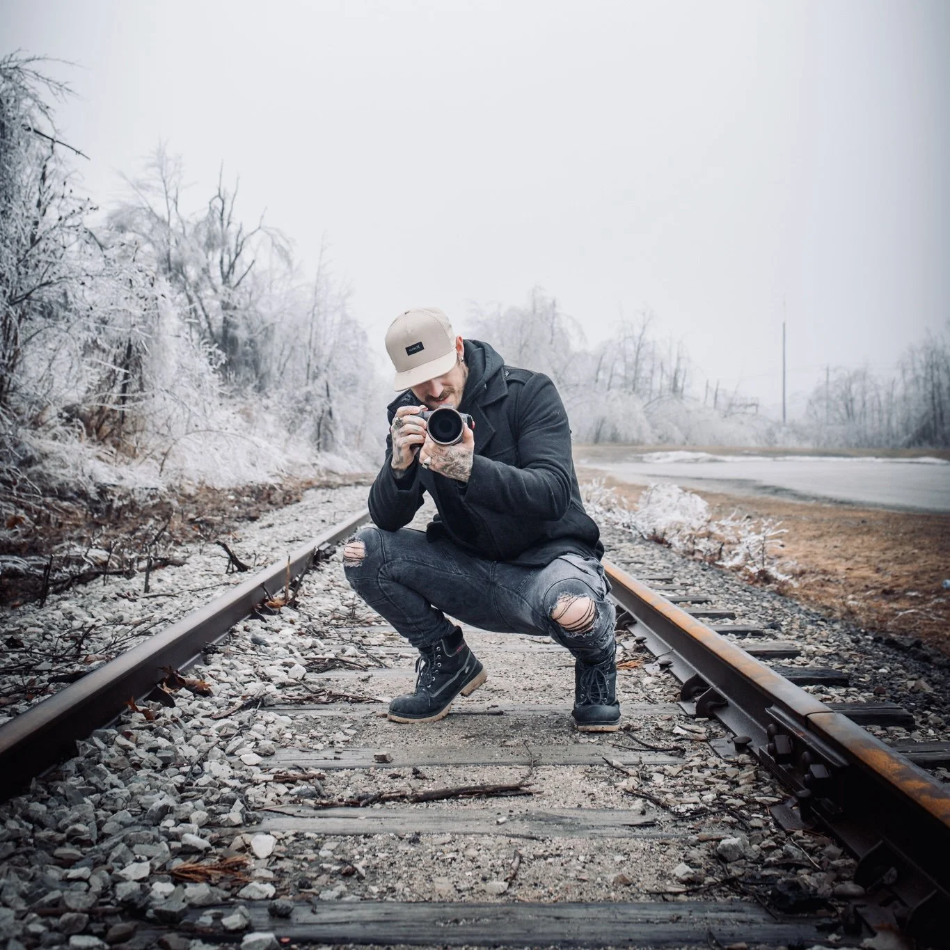 Man kneeling on train tracks taking a photo with camera in cold, snowy outdoor setting.
