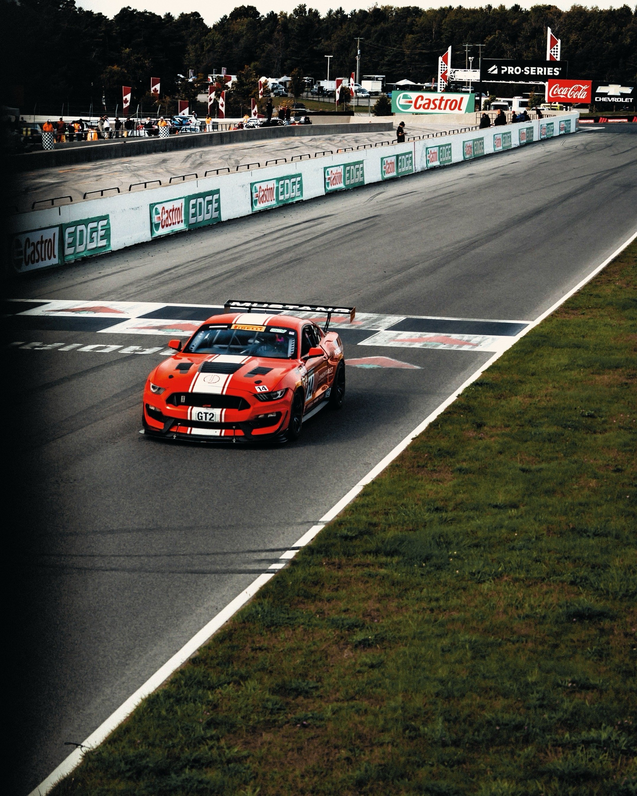 A red race car with black and white stripes crossing the finish line at a race track, with spectators and pit crews in the background.
