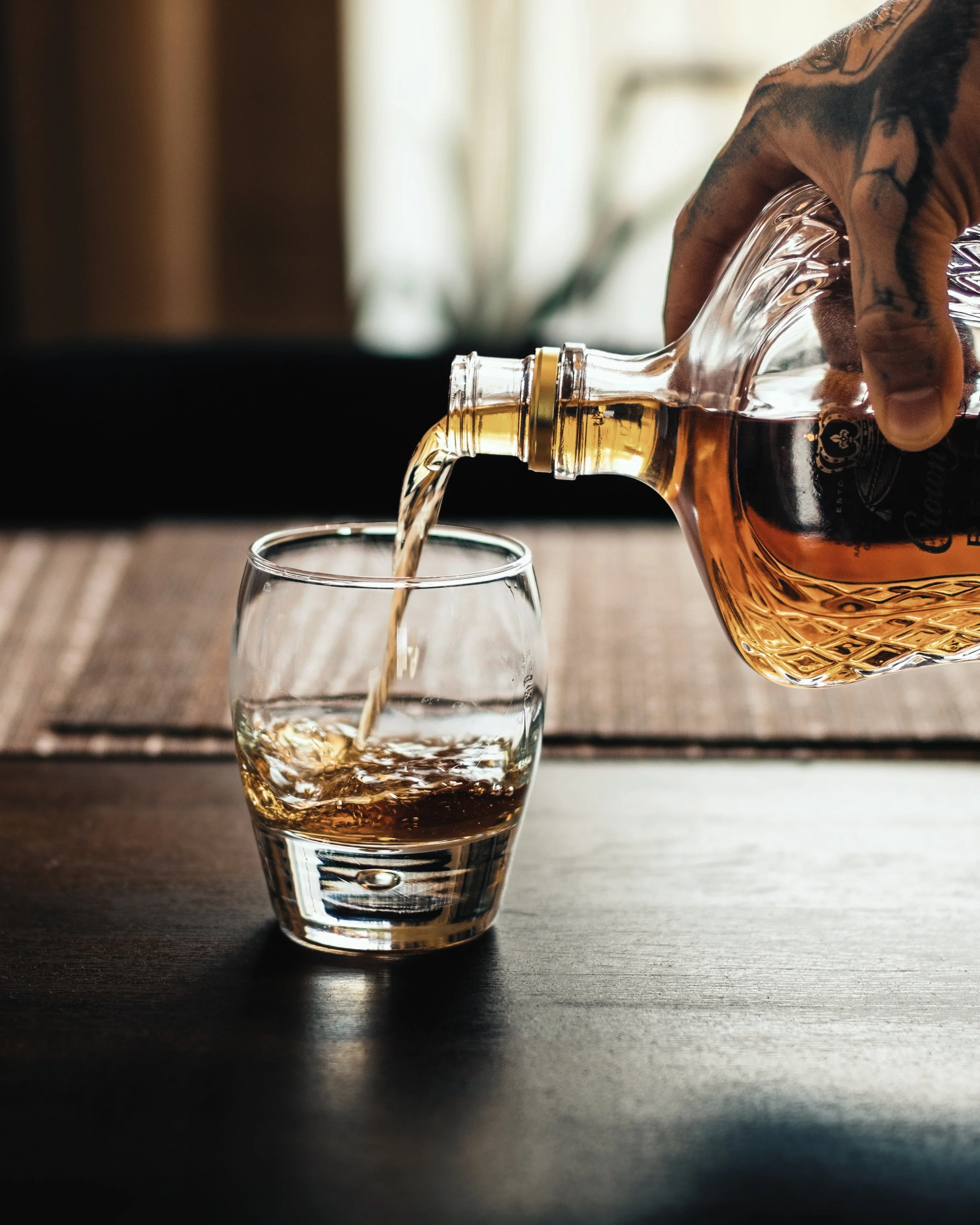 A hand with tattoos pouring whiskey from a glass bottle into a short glass on a dark wooden surface.