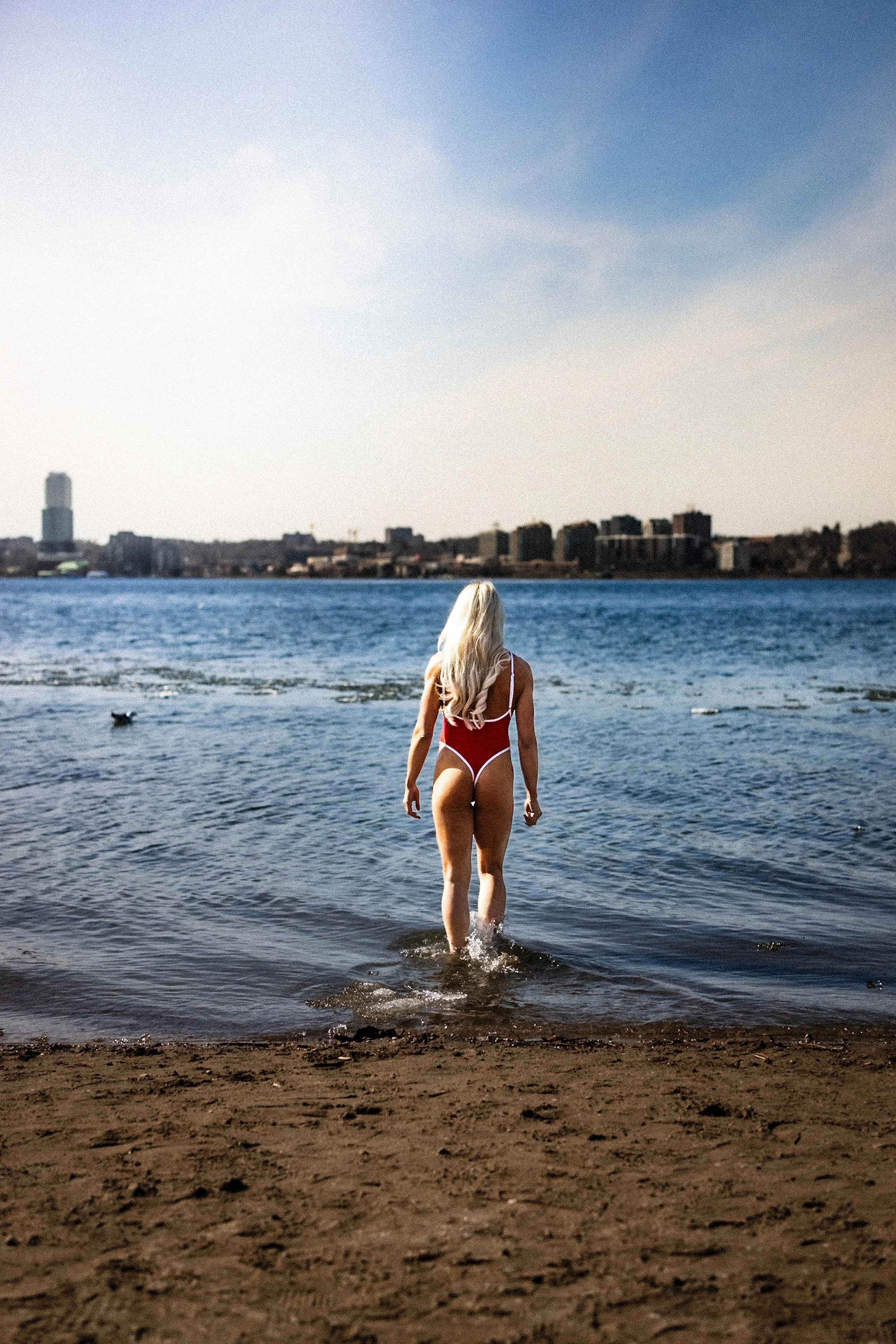 A woman in a red swimsuit walking into the water at the beach, with city buildings visible across the water and the sky partly cloudy.