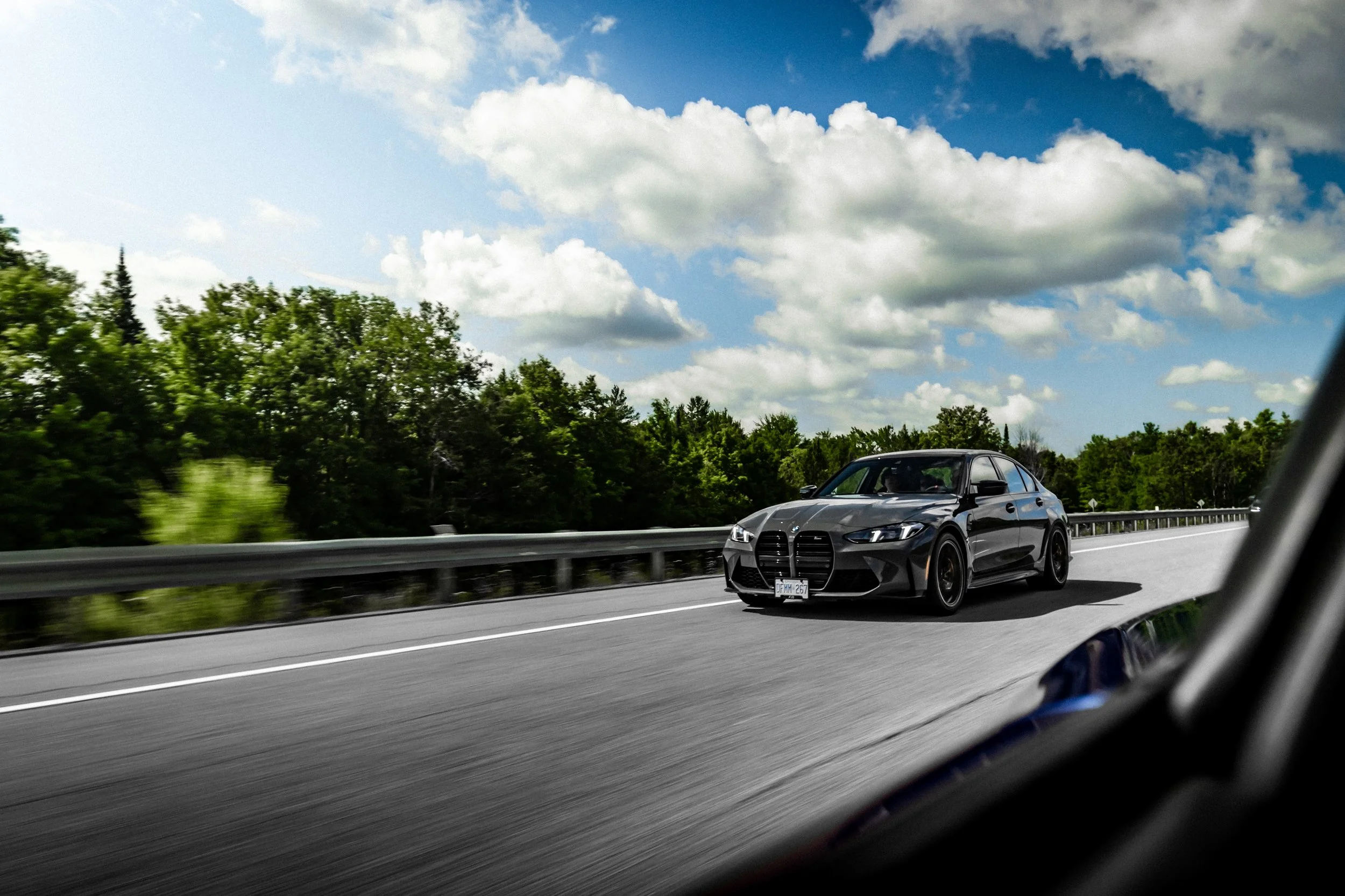 A black sports car driving along a highway with a guardrail, green trees, and a partly cloudy blue sky in the background, viewed from inside another vehicle.